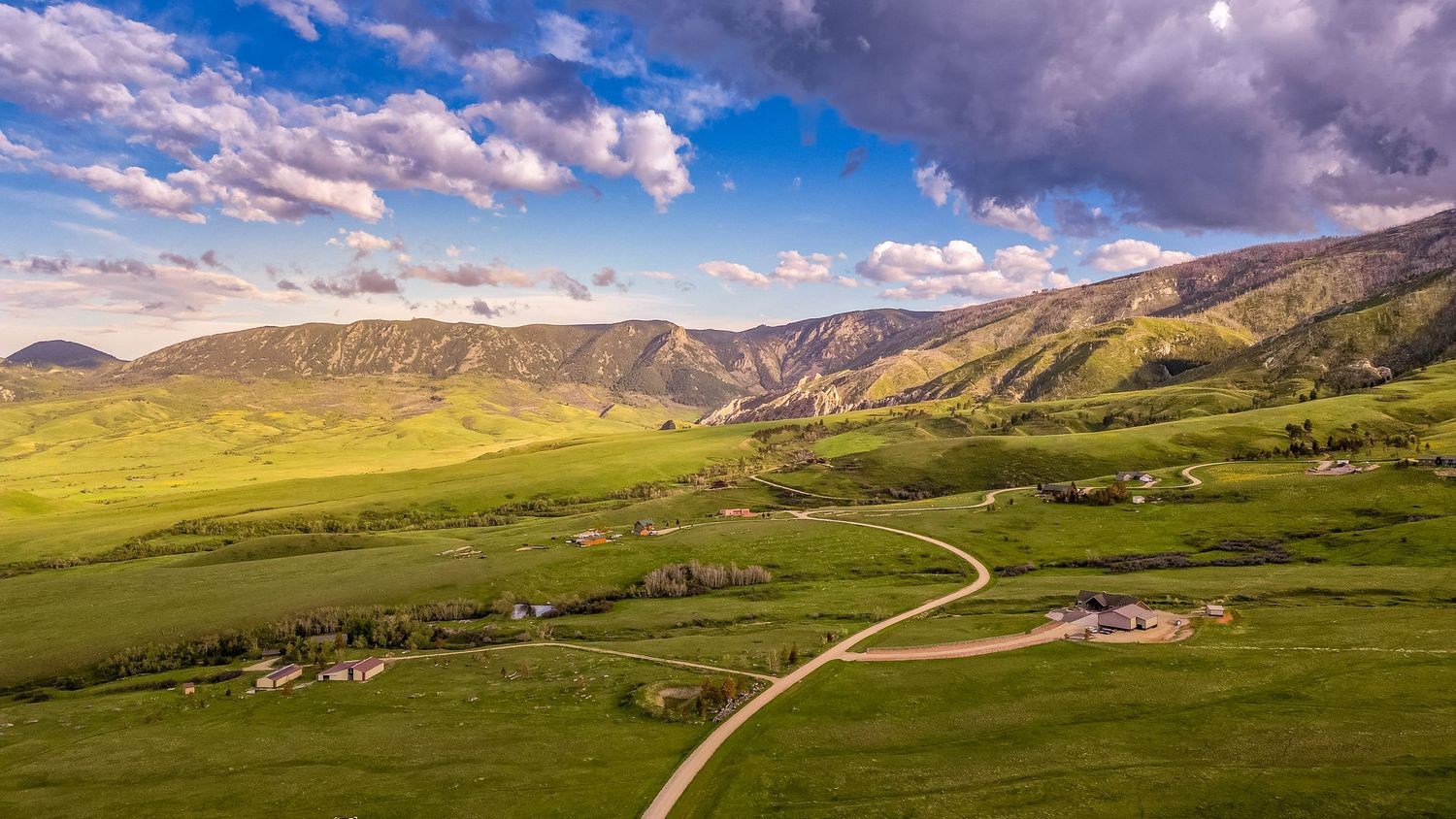 Aerial panoramic view of a green mountain valley with winding road and farmhouse.