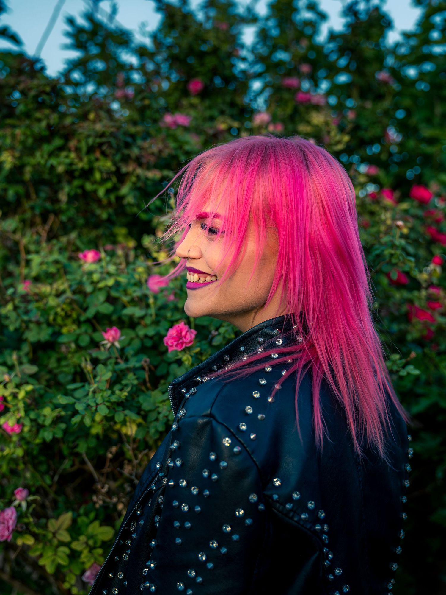 Pink-haired woman in studded leather jacket laughing outdoors near rose bushes.