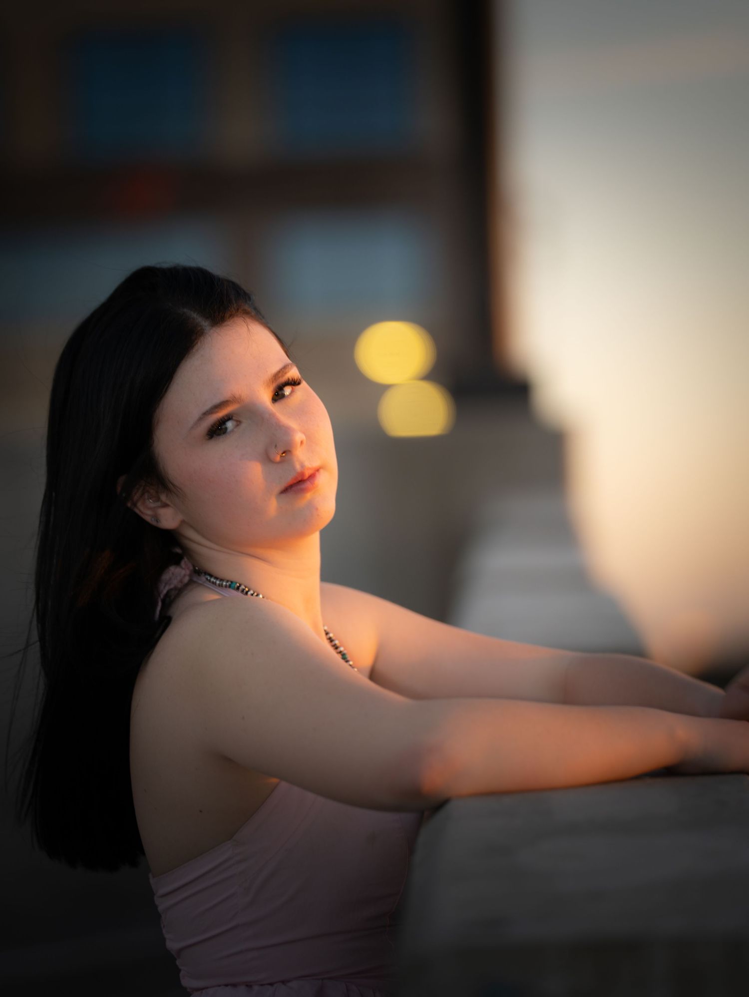Dark-haired young woman in pink dress poses at dusk with bokeh lights.