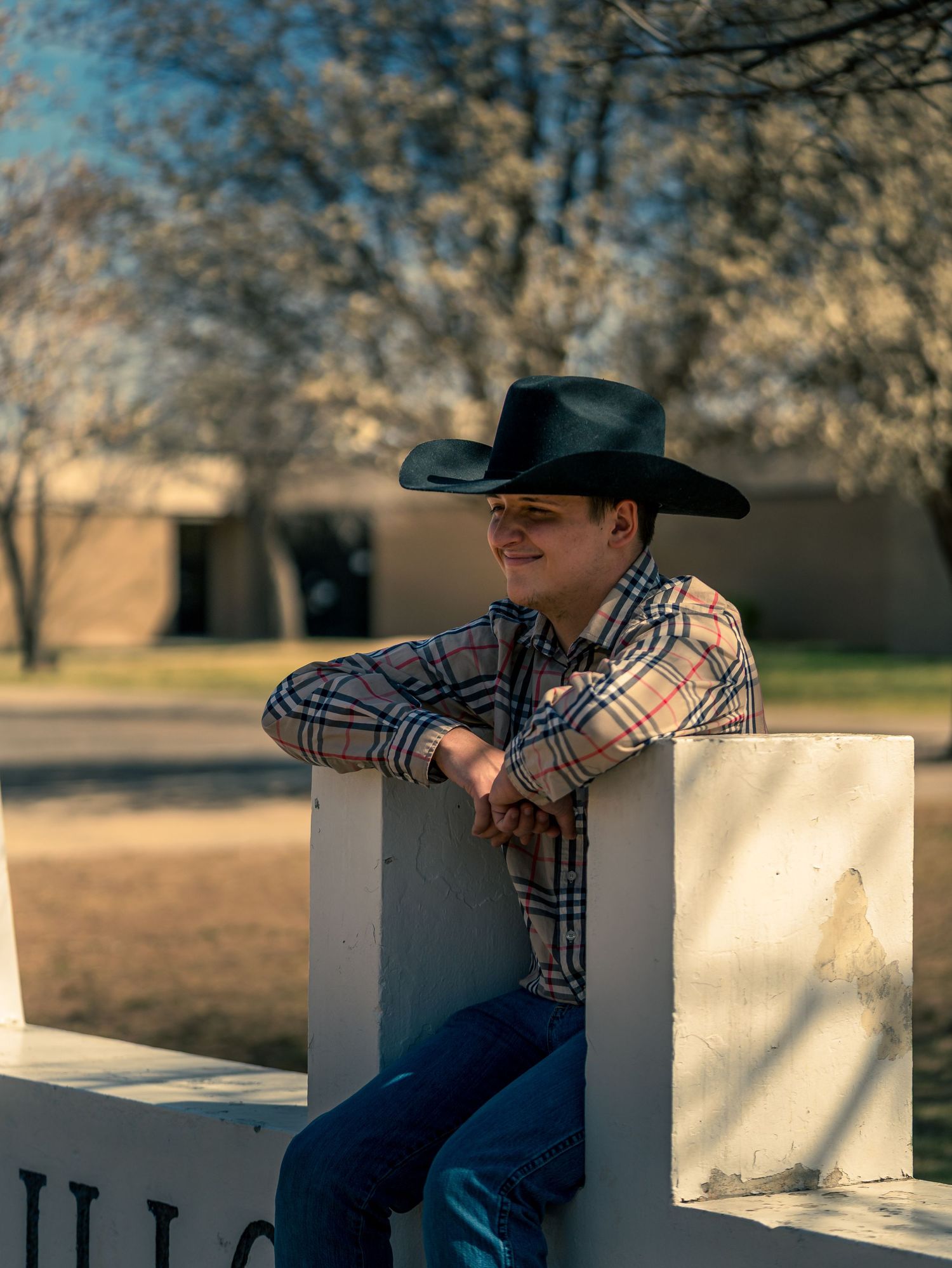 A smiling young boy wearing a black cowboy hat and plaid shirt sits relaxed on a white outdoor monument sign.