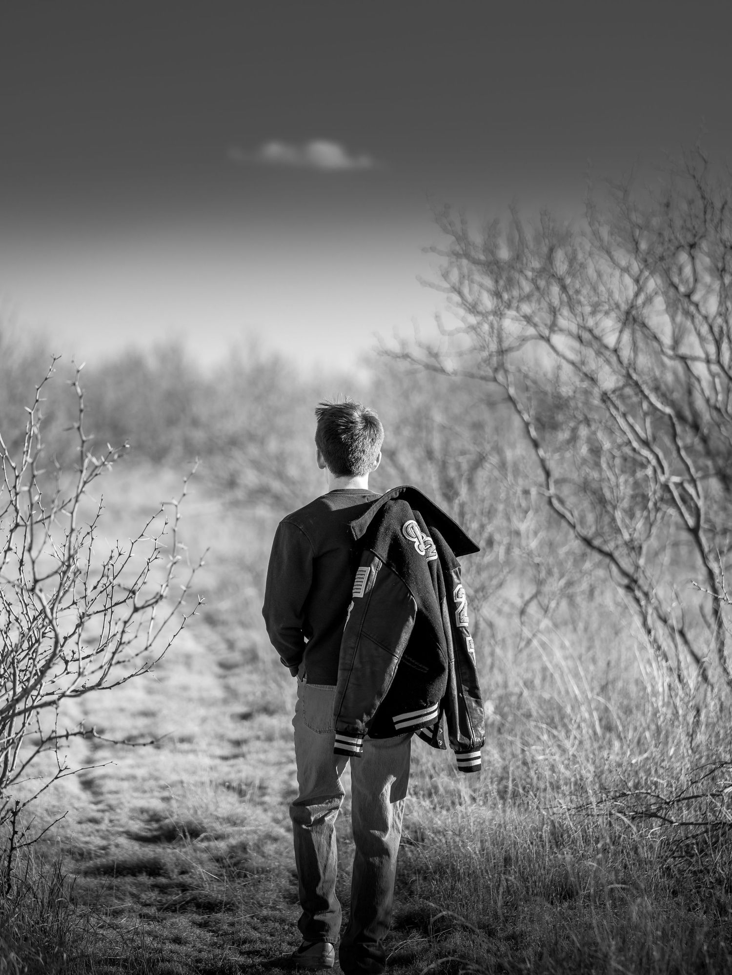 Black and white photo of a young person walking a rural path carrying a letterman jacket.