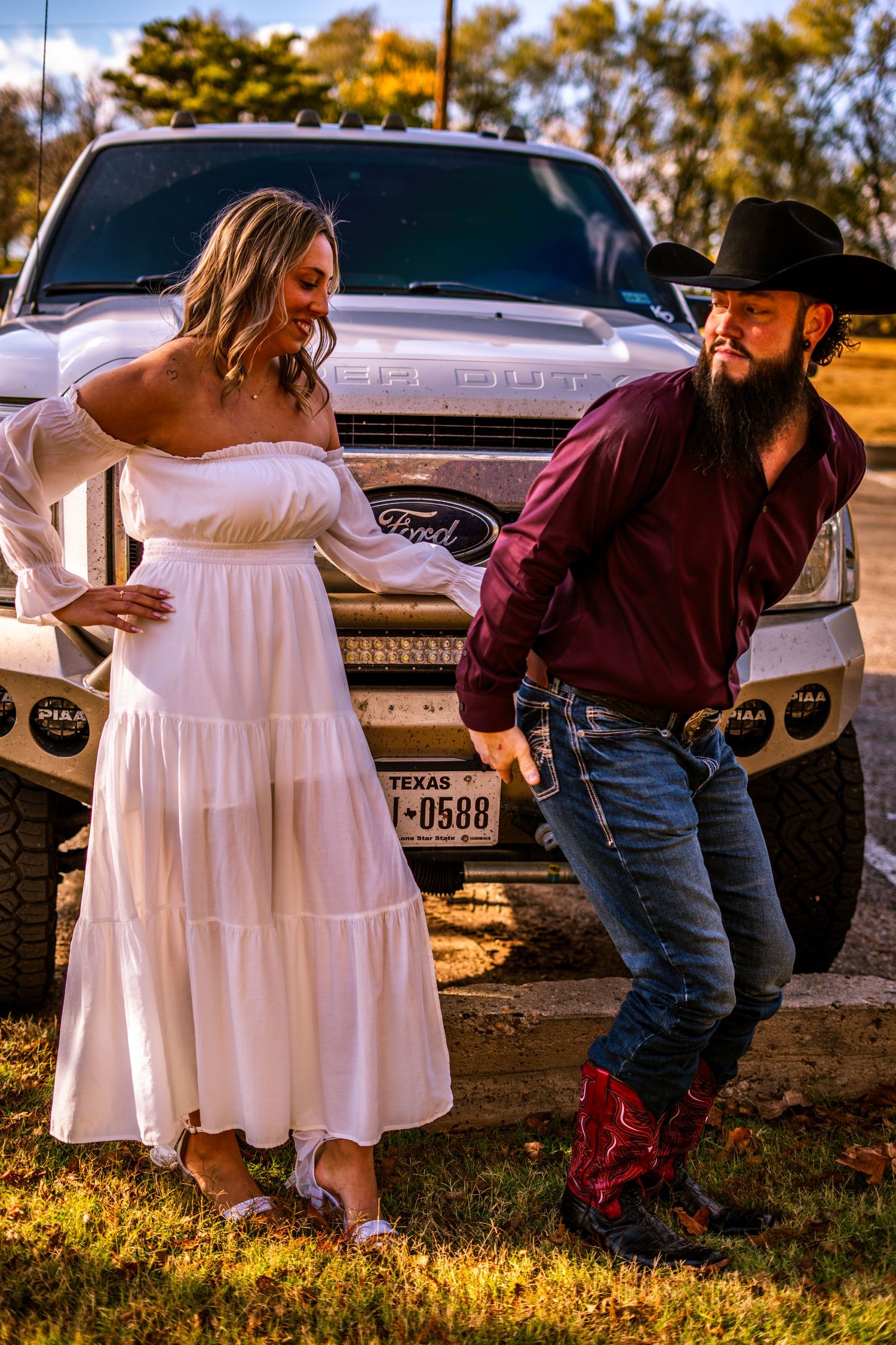 Couple posing in front of a Ford F-250 Texas truck, woman in white dress touching belly, man in cowboy hat.