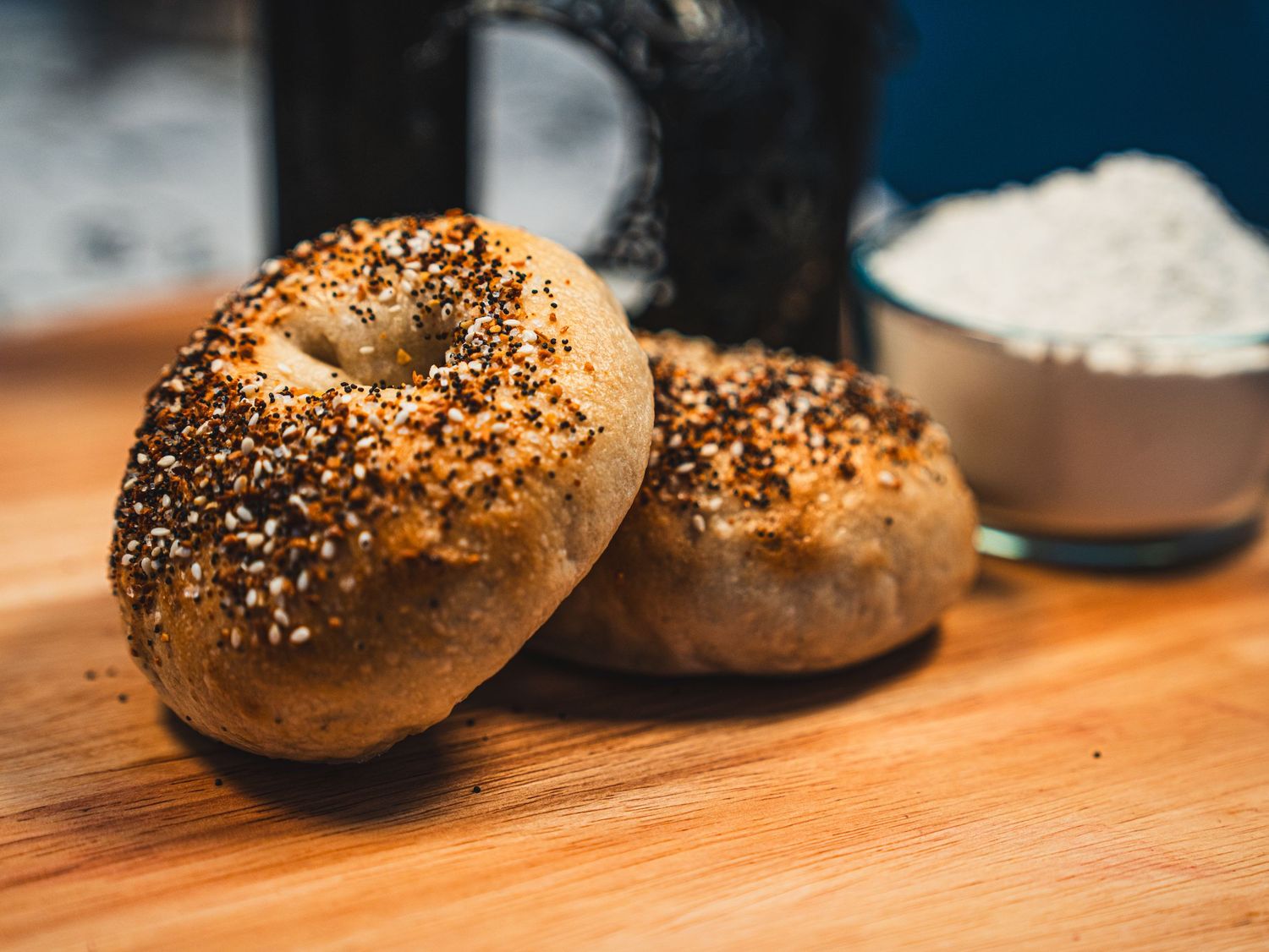 Two everything bagels topped with sesame and poppy seeds on a wooden cutting board with flour in background.