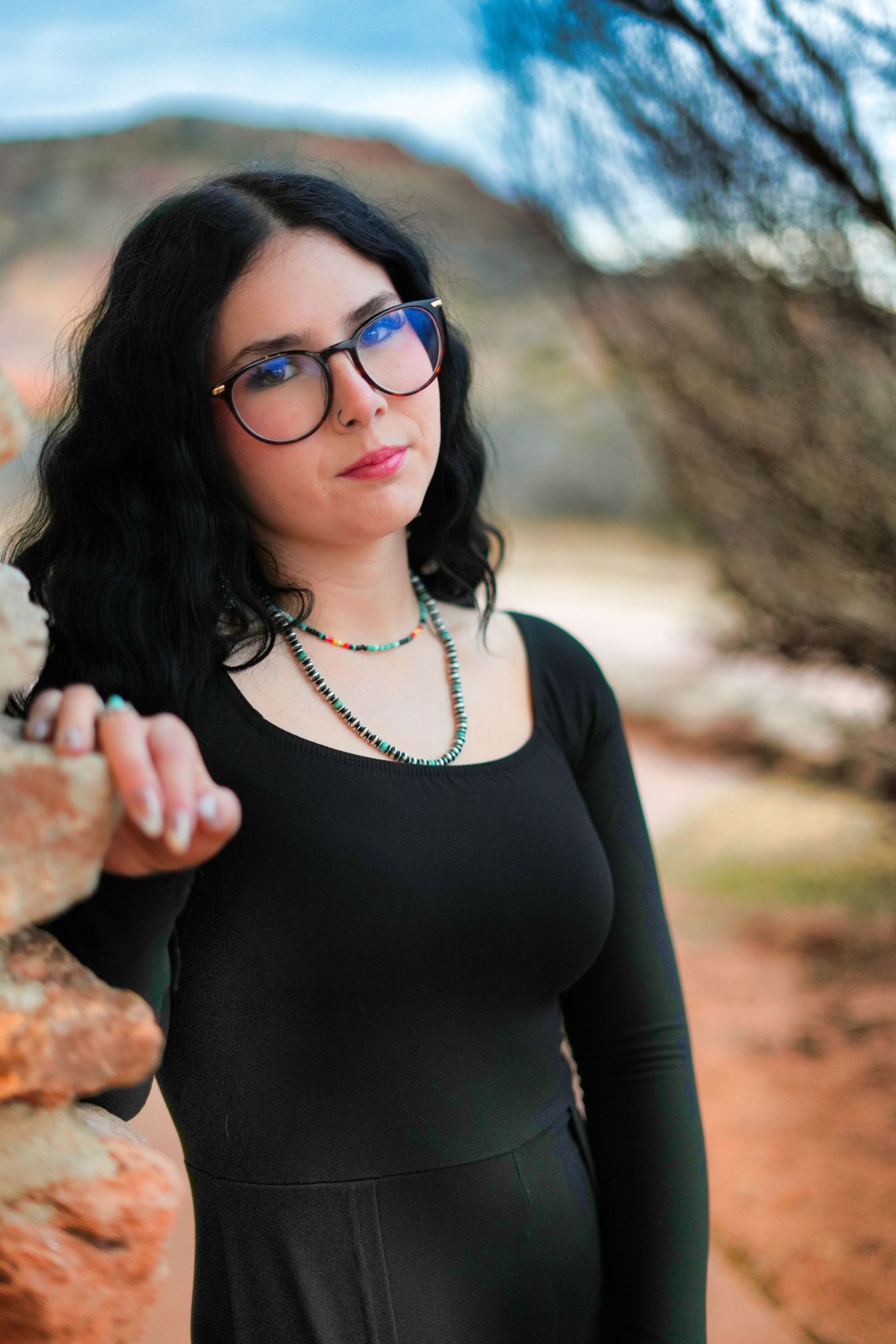 Young woman with black hair and glasses wearing a black dress poses outdoors near red rocks.