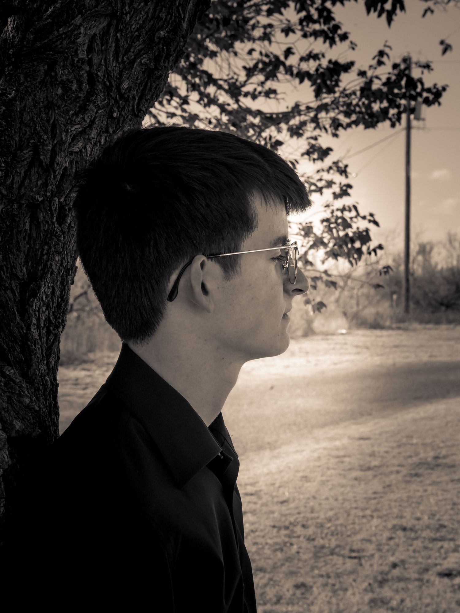 Black and white portrait of a young man with glasses leaning against a tree outdoors.