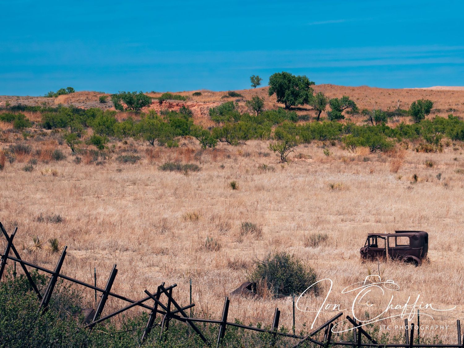 An abandoned rusty vehicle sits in a dry golden field beside a weathered wooden fence under a clear blue sky.