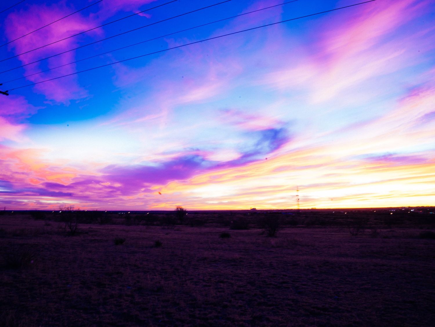 Vivid purple and gold sunset sky over a dark flat desert landscape with power lines.