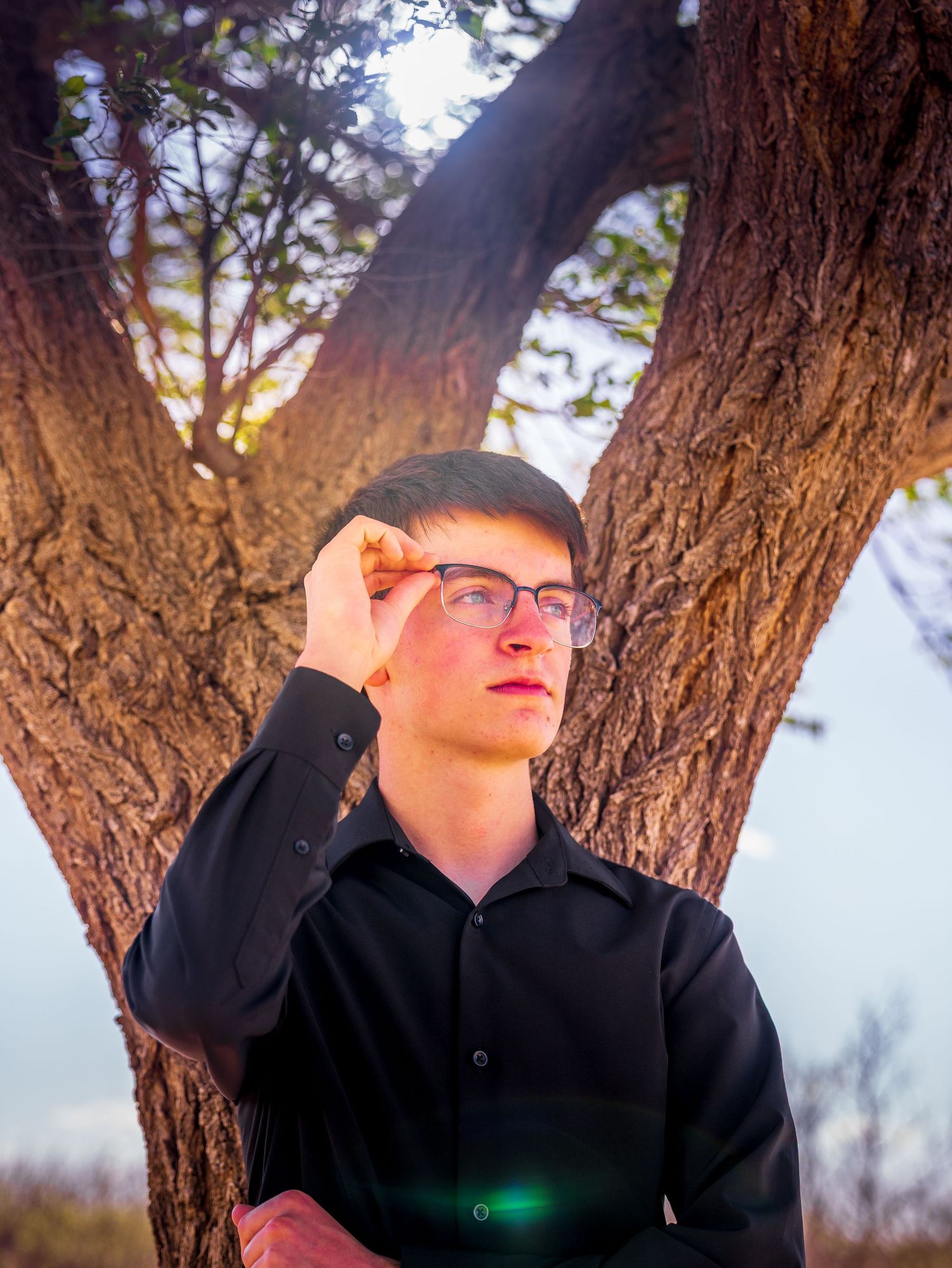Young man in black shirt adjusting glasses while standing in front of a textured tree trunk.