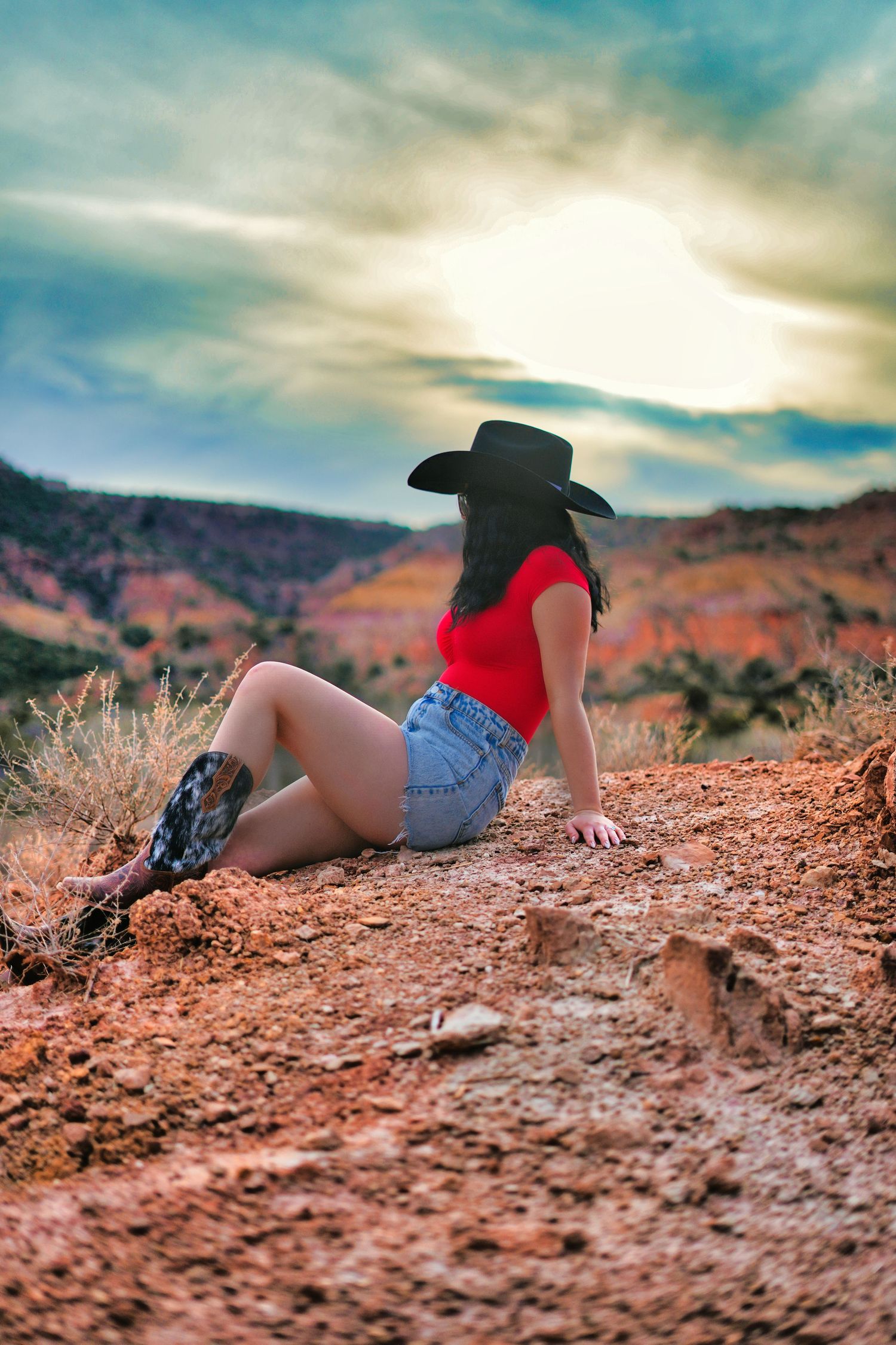 Woman in red top, denim skirt, and cowboy hat sits on red rock at dramatic desert sunset.