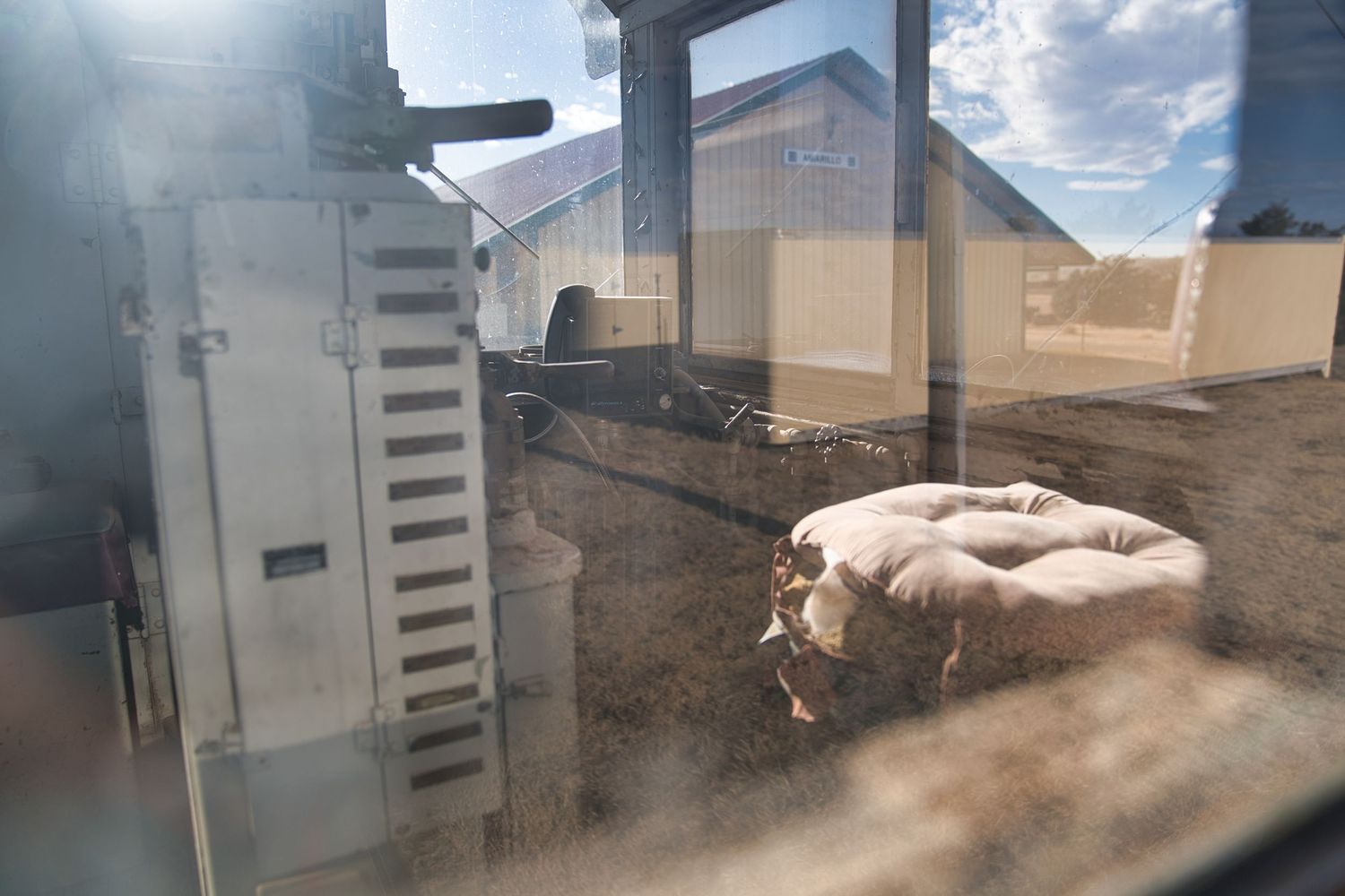 View through a dusty window of old farm equipment with burlap sacks and agricultural buildings in background.