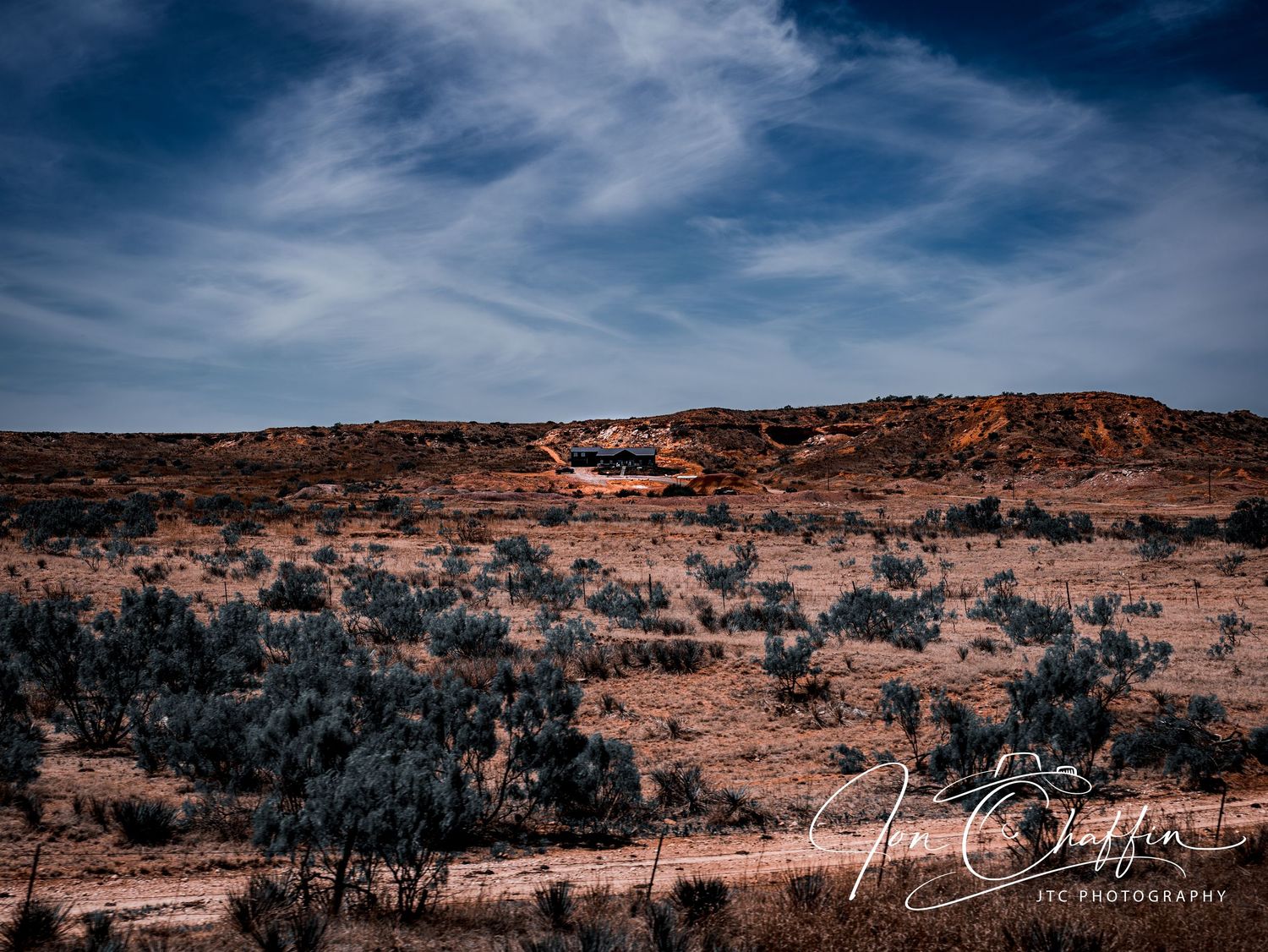A vast arid desert landscape with sparse shrubs and a small structure on a red mesa under a dramatic blue sky.