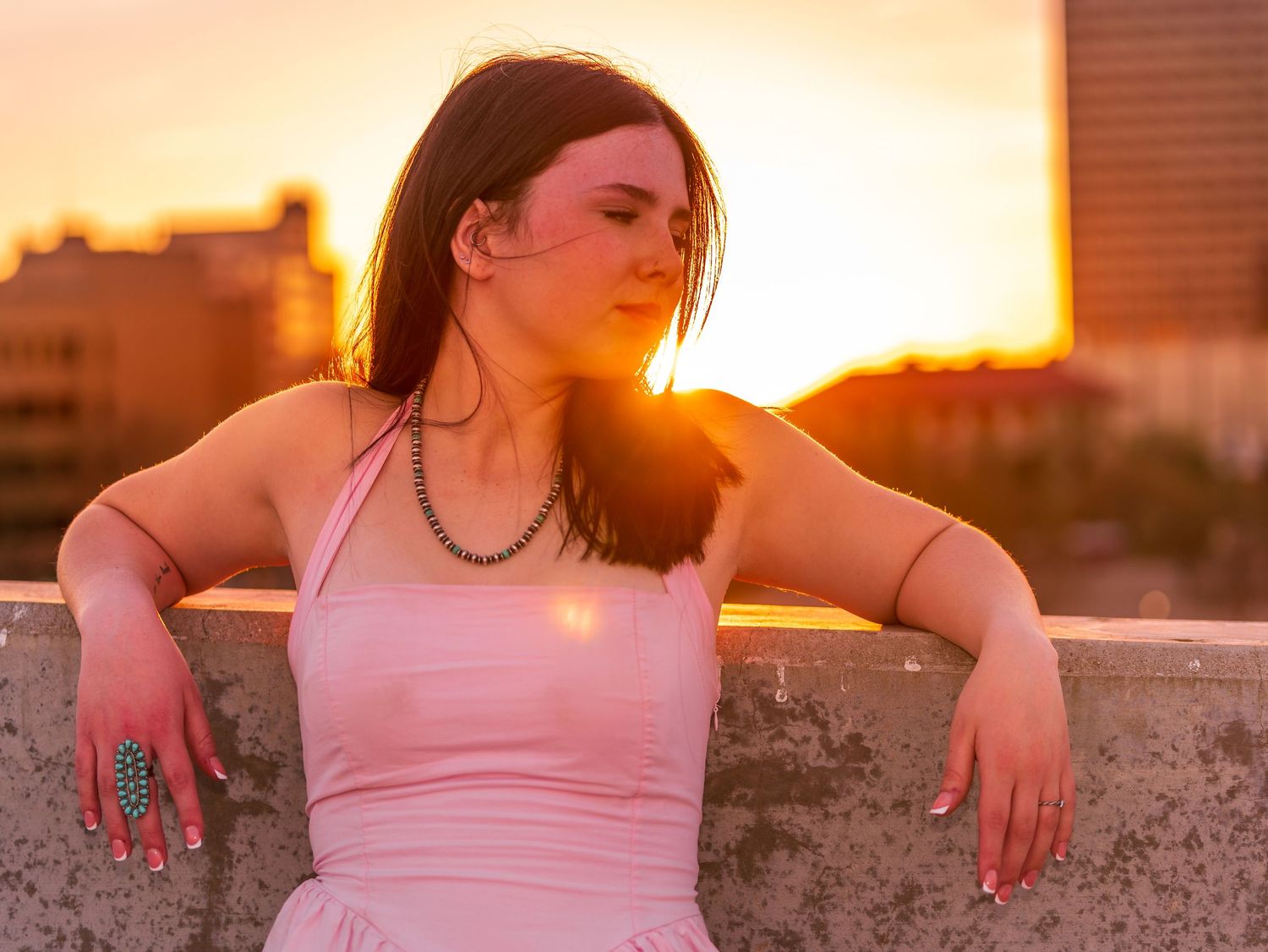 Young woman in pink dress poses on rooftop during vibrant sunset.
