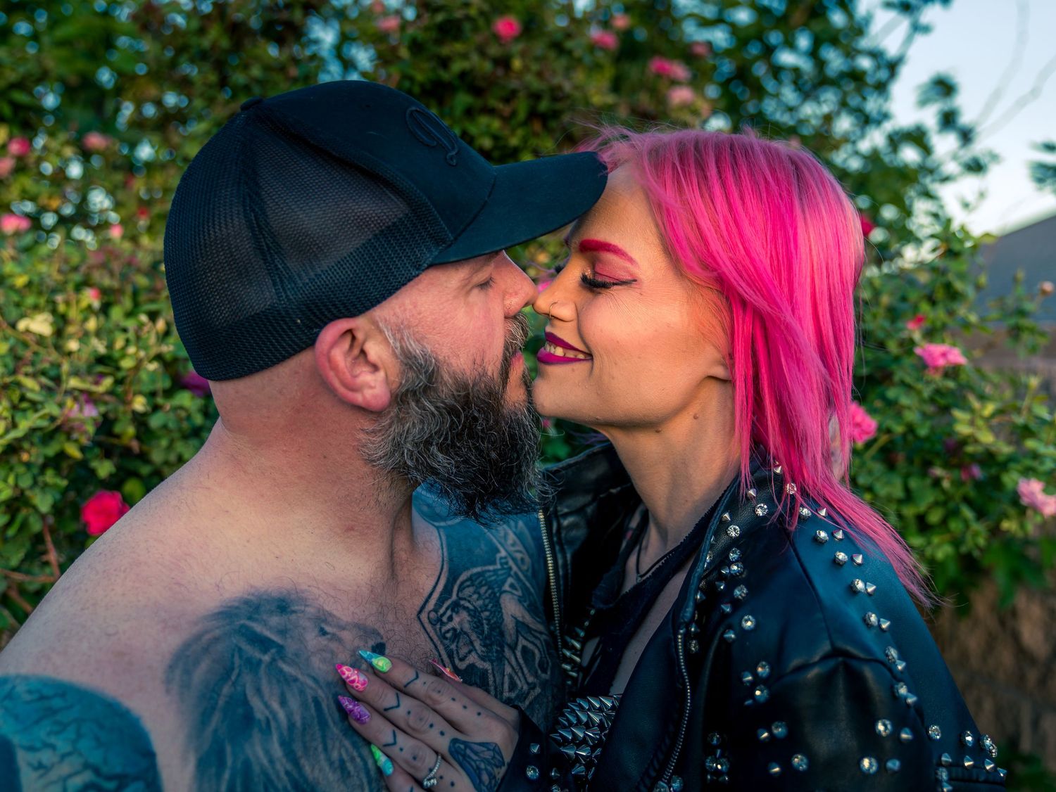 Couple sharing a romantic kiss outdoors, woman with vibrant pink hair wearing a studded leather jacket.