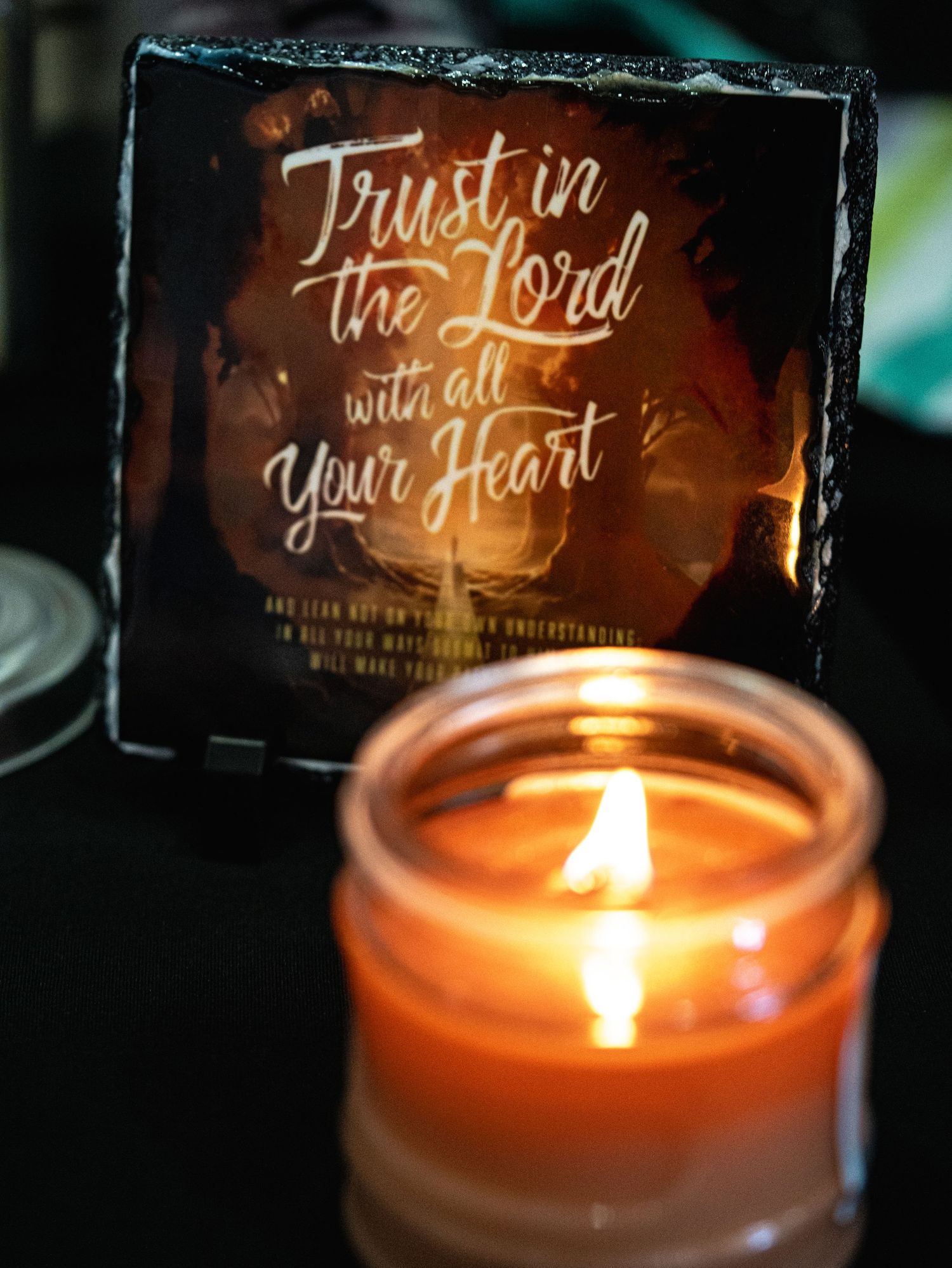 A glowing candle in a jar beside a slate tile with the Bible verse Trust in the Lord with All Your Heart.