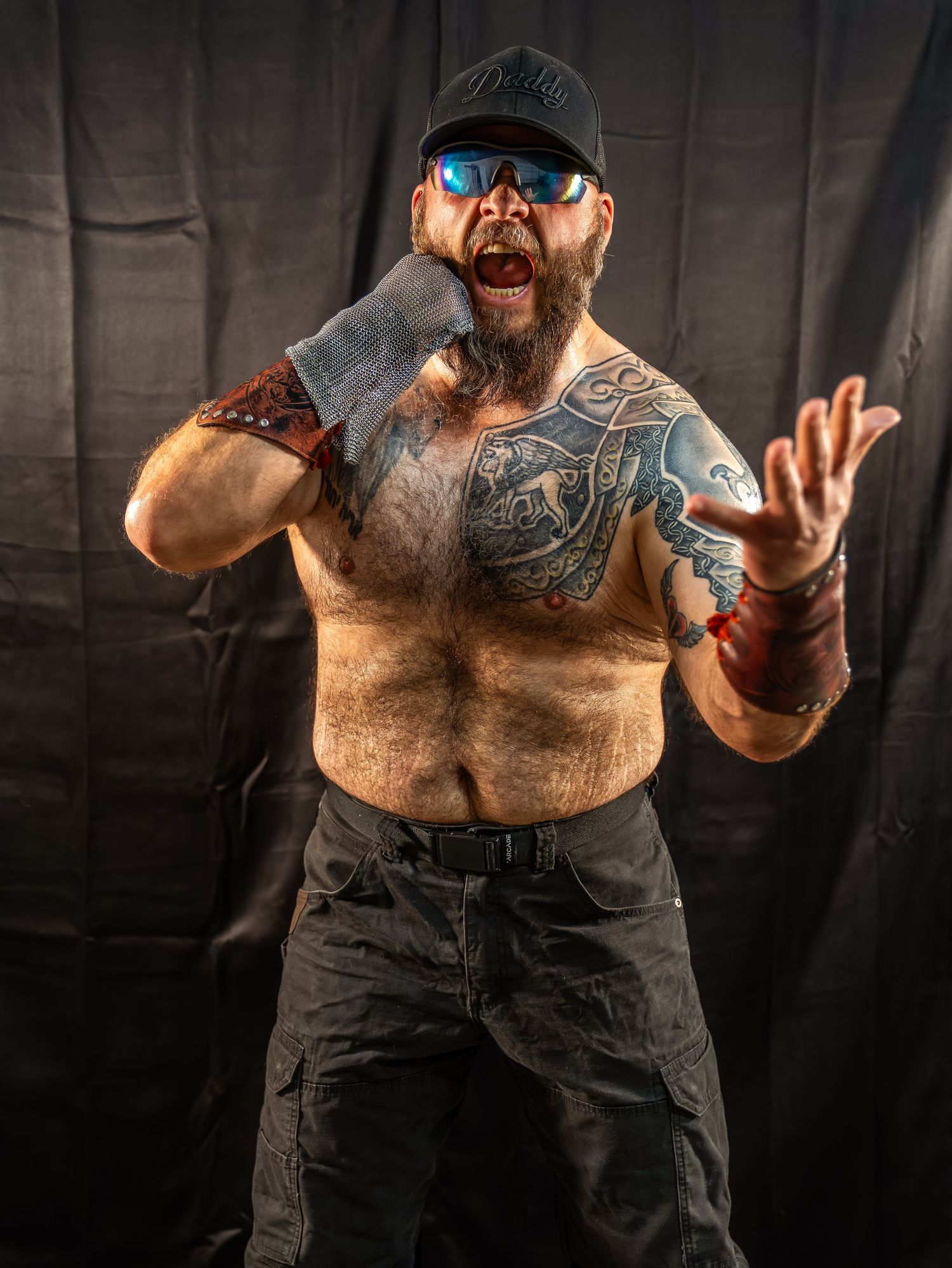 Shirtless tattooed man with beard wearing cap and sunglasses poses dramatically against black backdrop.