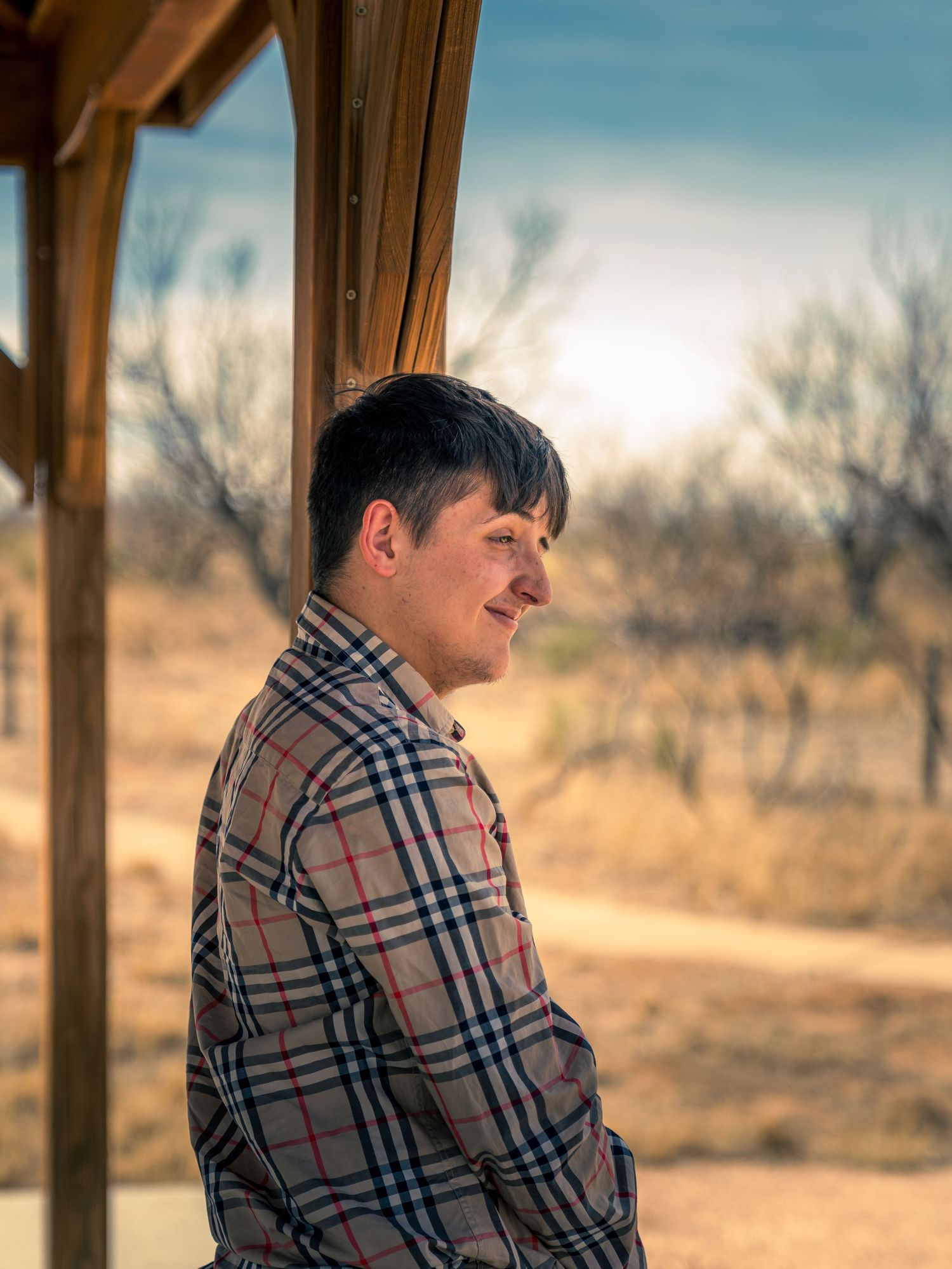 Young man in plaid shirt smiling in profile under a pergola with sunny rural backdrop.