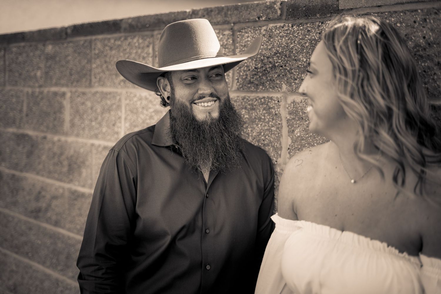 Sepia-toned portrait of bearded man in cowboy hat and woman in white dress smiling at each other.