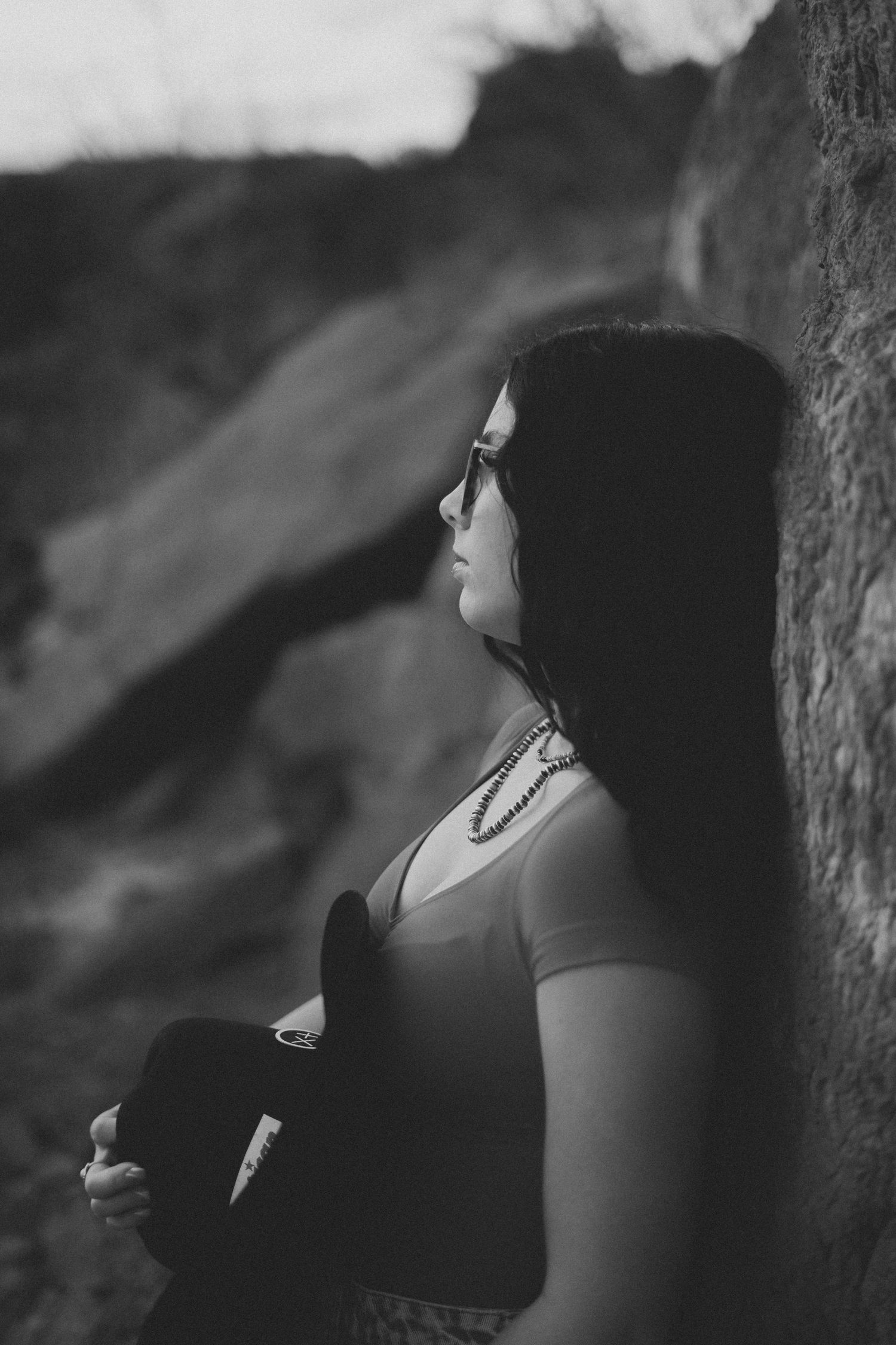 Black and white portrait of woman leaning against rock wall holding cowboy hat outdoors.