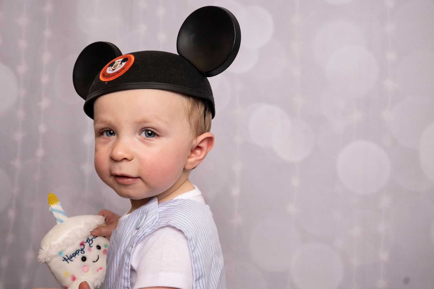 One-year-old wearing Mickey ears and holding a plush cupcake during a birthday milestone session by a children's Photographer