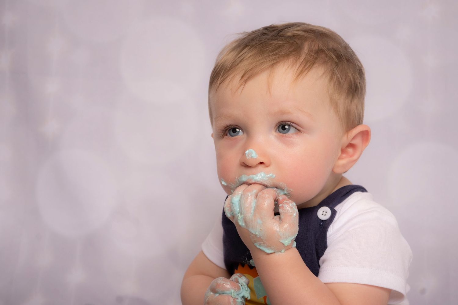 Toddler enjoying birthday cake with frosting-covered hands during a cake smash session by a Milestone Photographer.