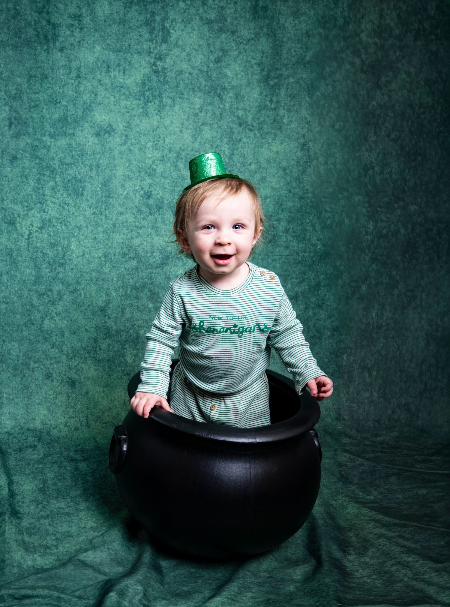 Smiling toddler sitting in a black bucket against a teal backdrop during a milestone portrait by a Central Florida Photographer.