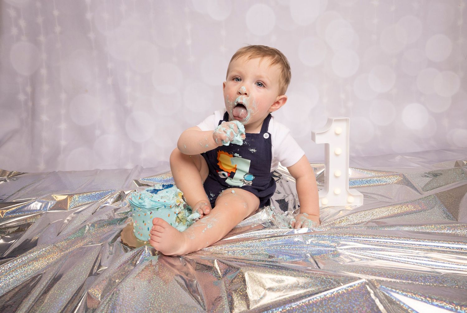 One-year-old beside a blue birthday cake and number one sign during a cake smash session by a Central Florida Photographer