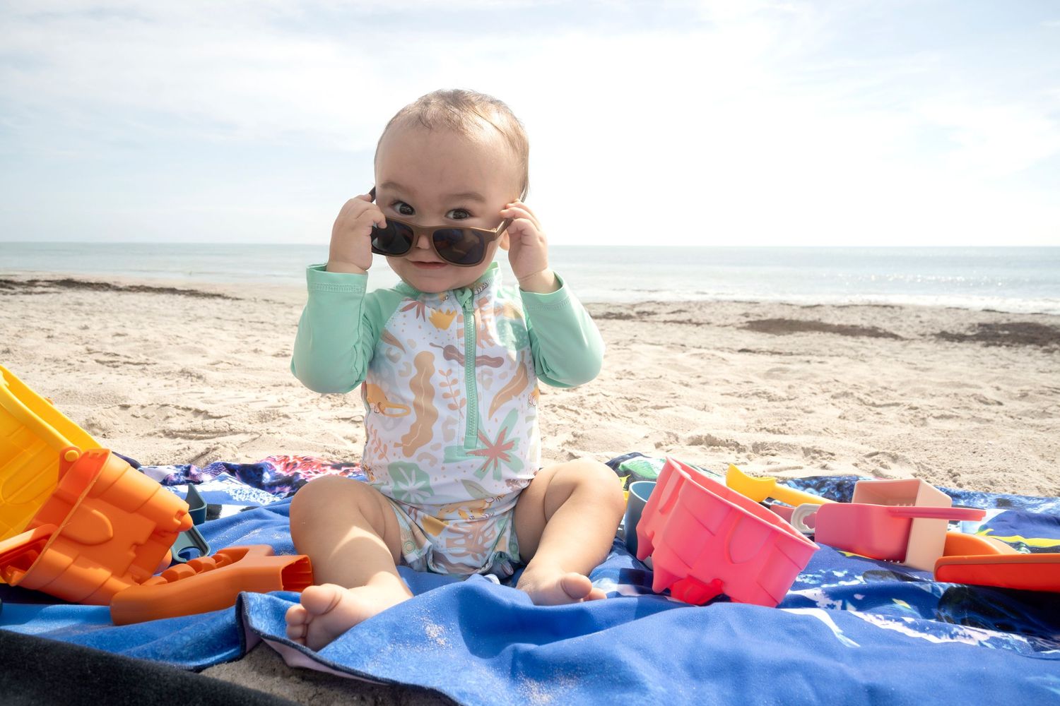 Baby wearing sunglasses while sitting on a beach blanket during a milestone session by a Central Florida Photographer.