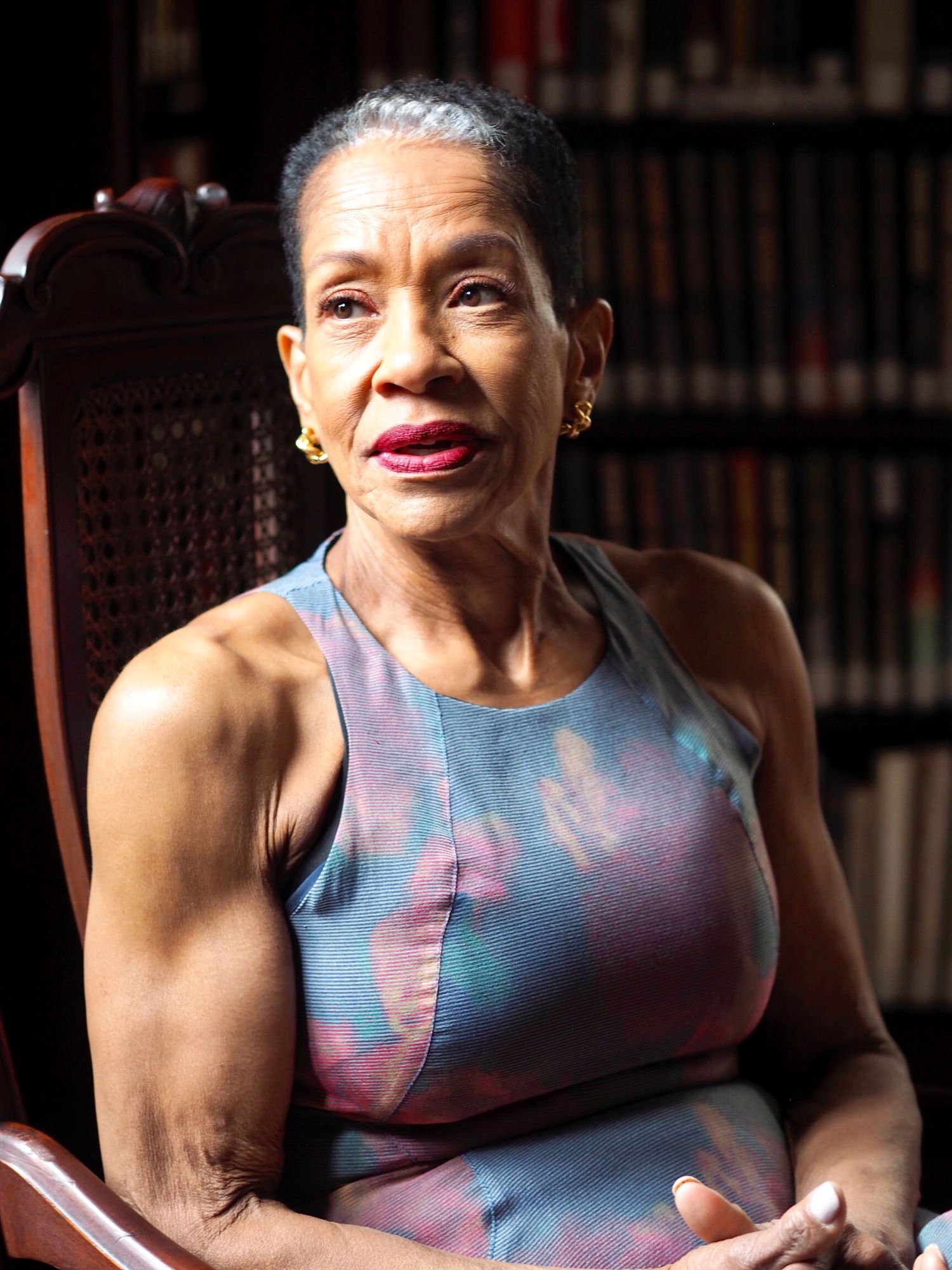 An elegant older Black woman sits in a wooden chair wearing a colorful sleeveless top, with bookshelves behind her.