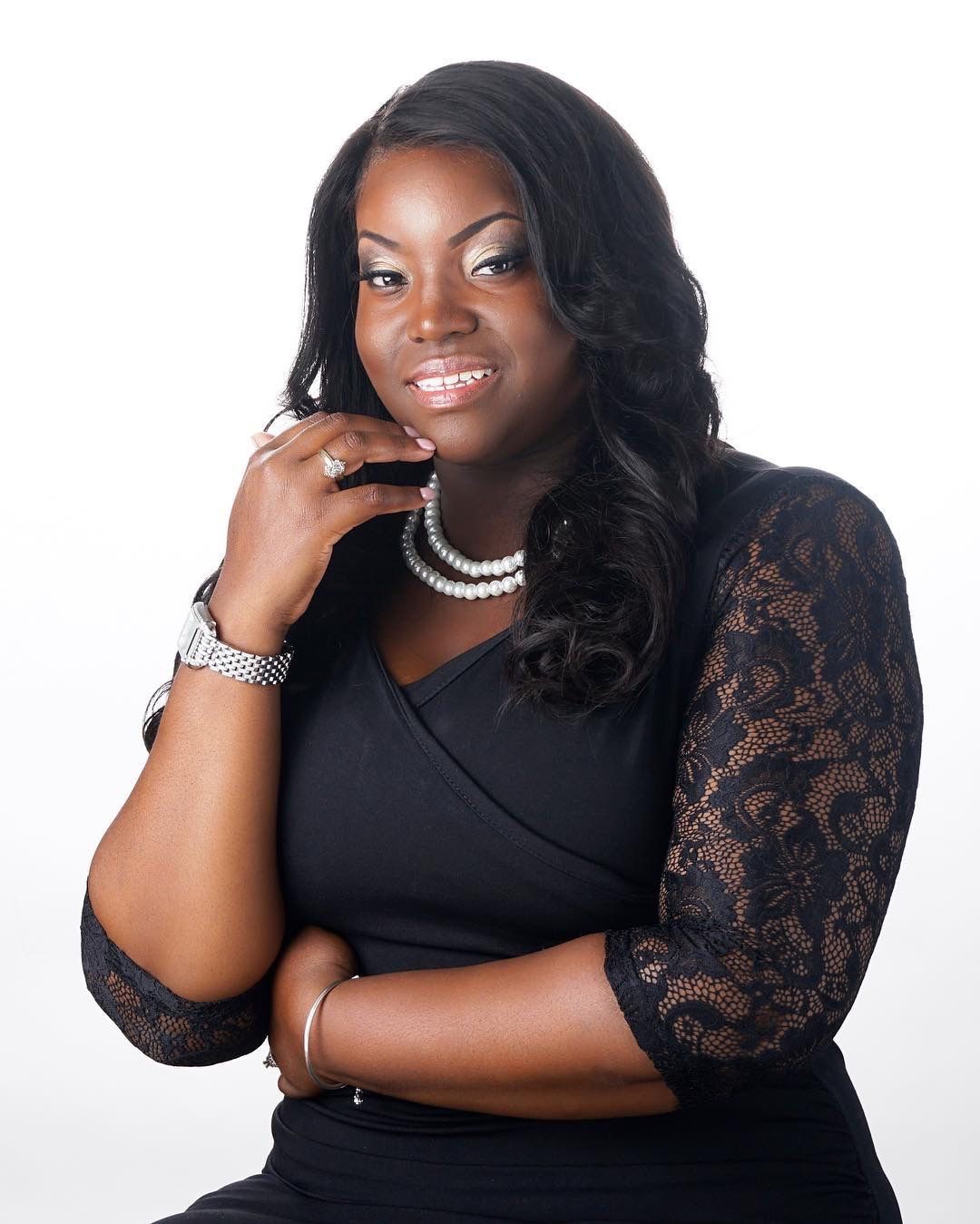 Smiling woman in a black lace dress with pearl necklace posing for a professional portrait.
