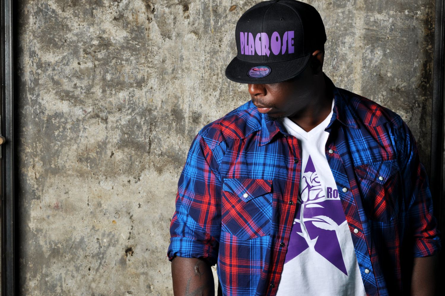 Young man in a plaid shirt and snapback cap posing against a weathered outdoor wall.