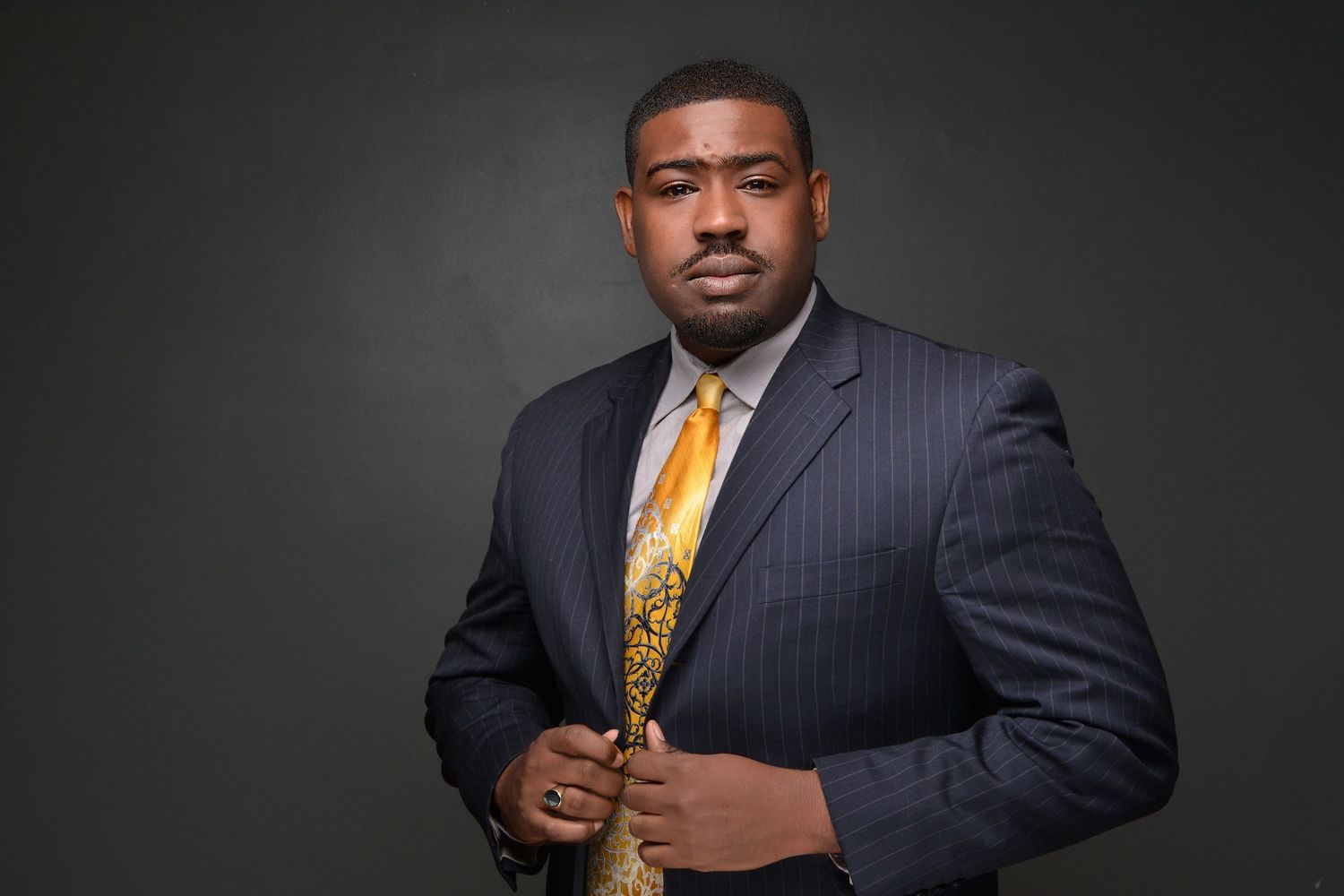 Professional Black man in navy pinstripe suit with gold tie posing confidently in studio.