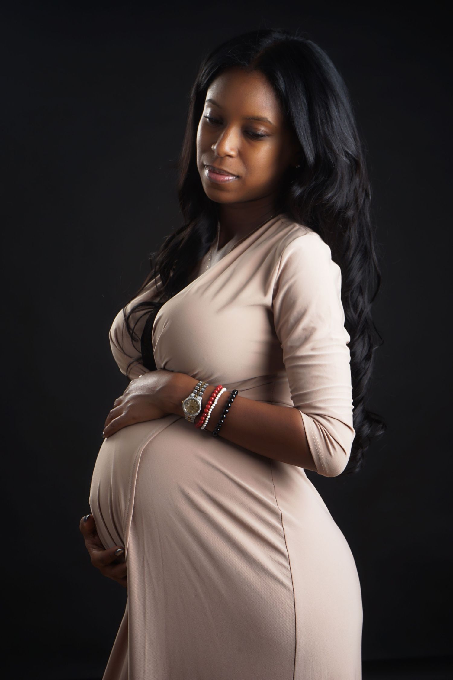 Elegant pregnant woman in beige dress cradles baby bump against dark studio background.