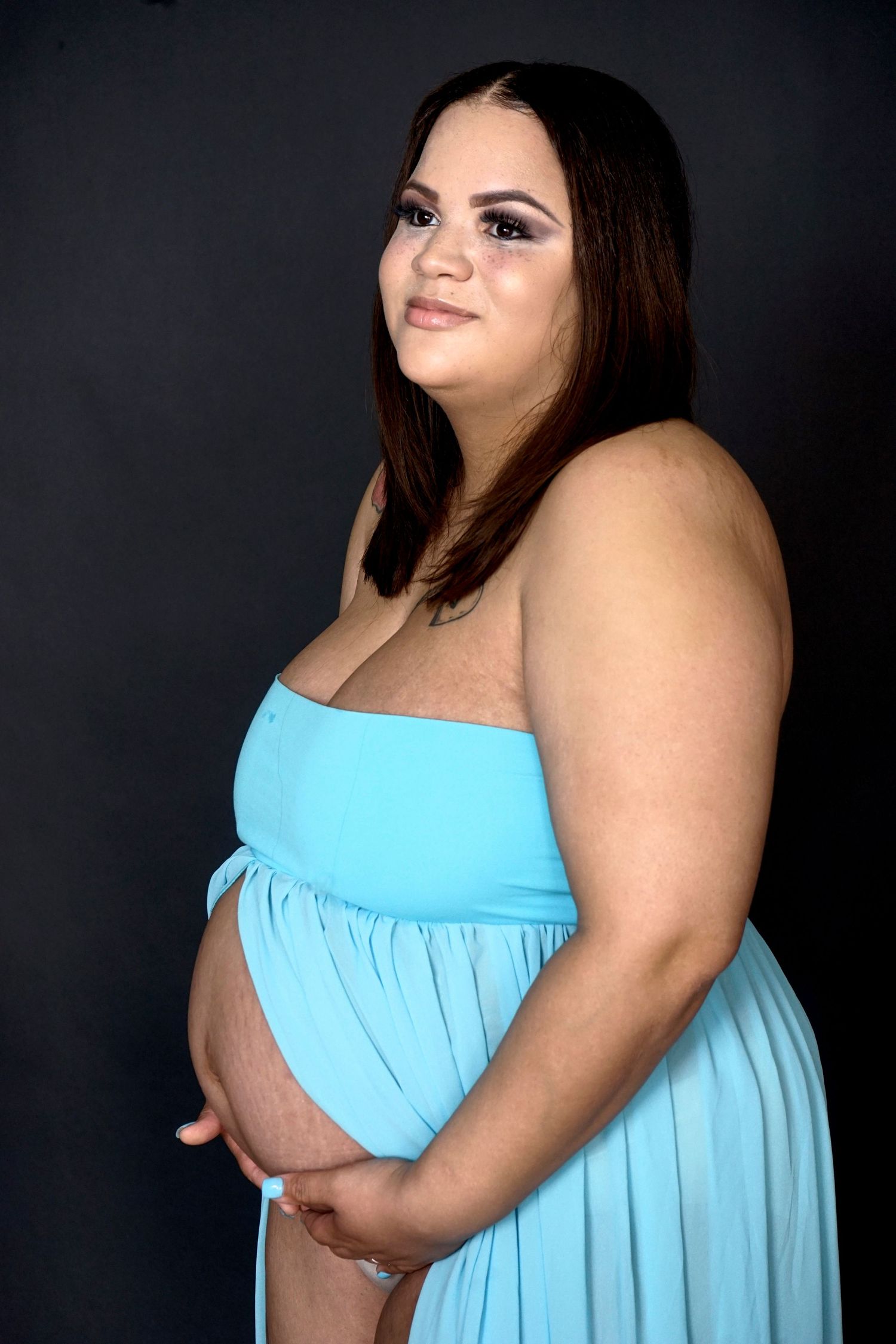 Plus size pregnant woman in blue strapless maternity dress holding baby bump against dark background.