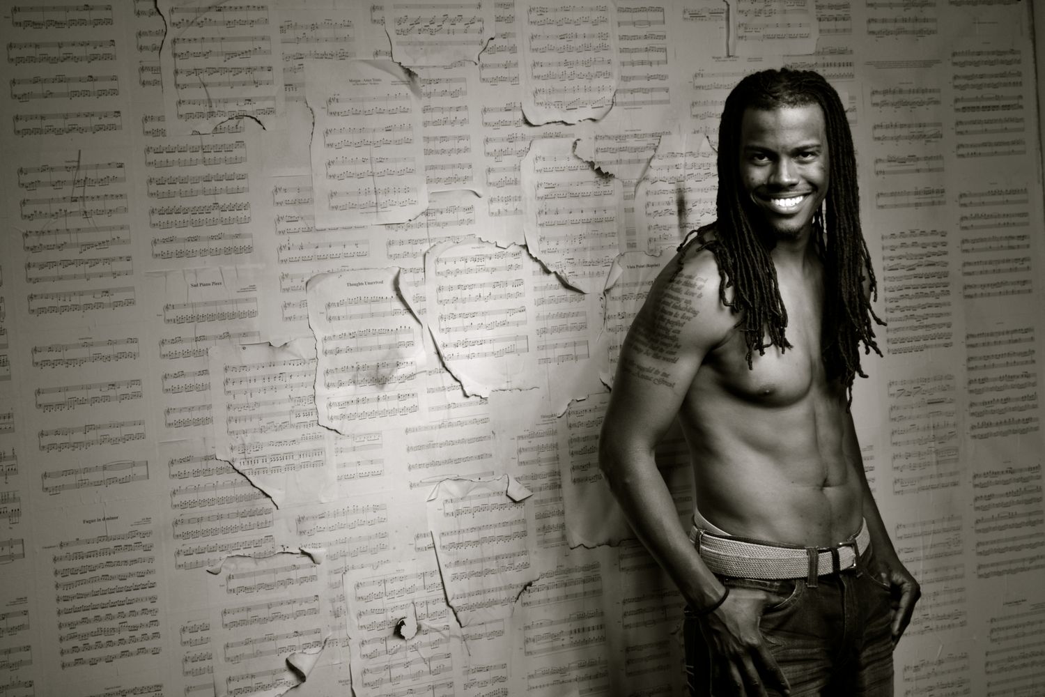 A shirtless smiling man with dreadlocks leans against a wall covered in torn sheet music pages.