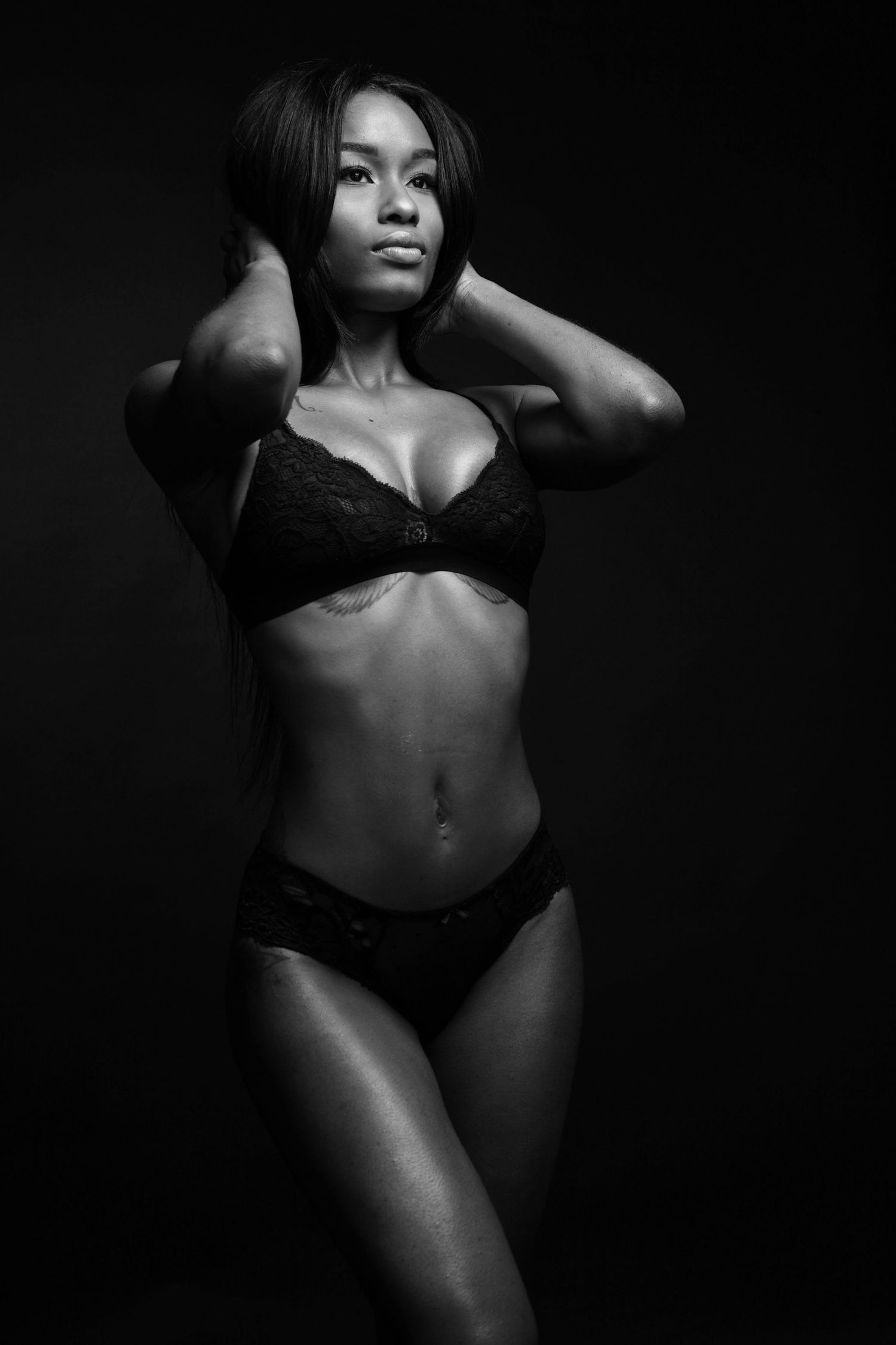 Black and white photo of a fit Black woman posing confidently in black lace lingerie against a dark background.