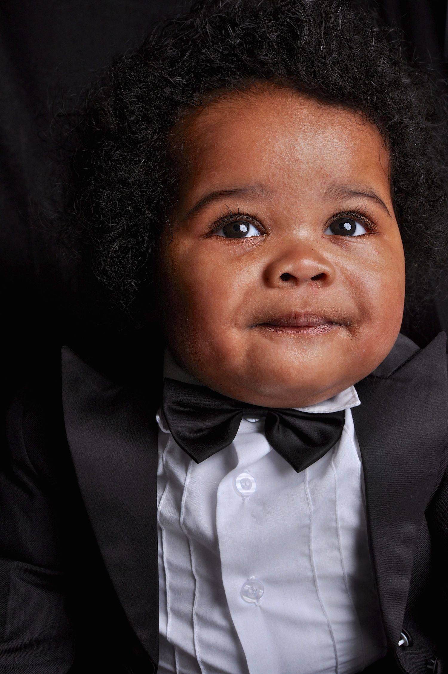 Adorable Black baby boy dressed in a formal black tuxedo with bow tie posing for a portrait.