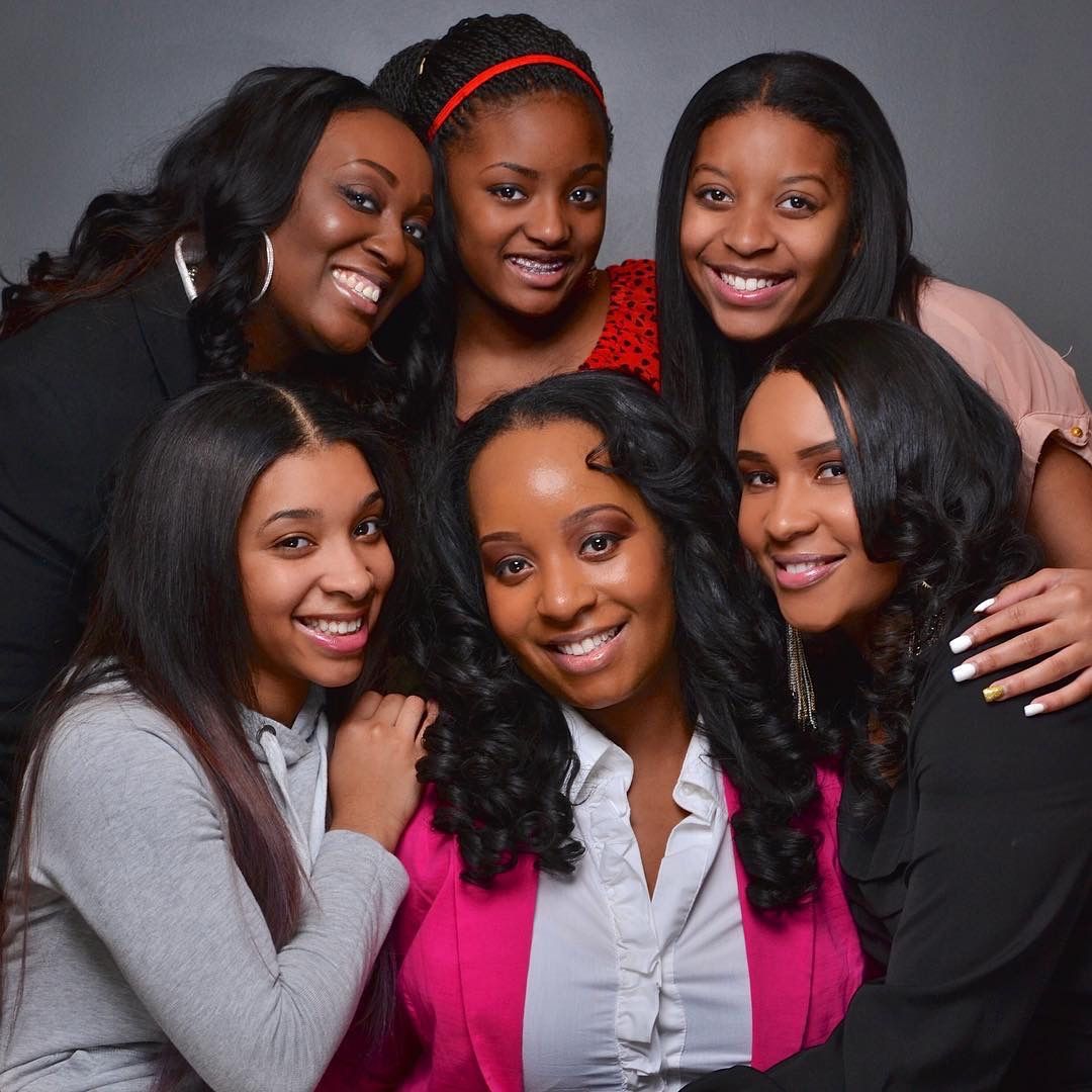 Six young women pose together smiling in a professional group portrait against a gray studio background.