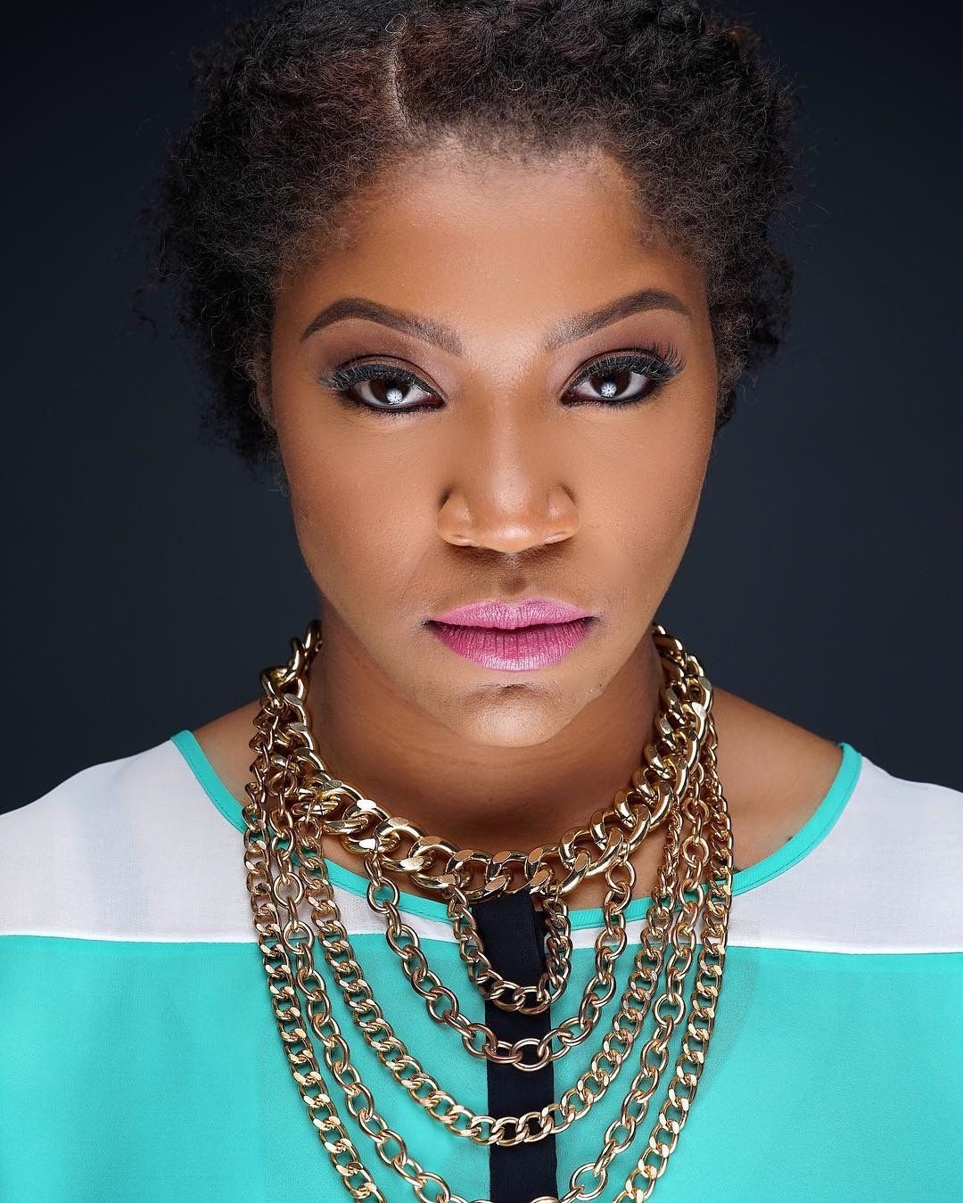 Bold close-up portrait of woman with natural hair wearing gold chain necklace and teal top.