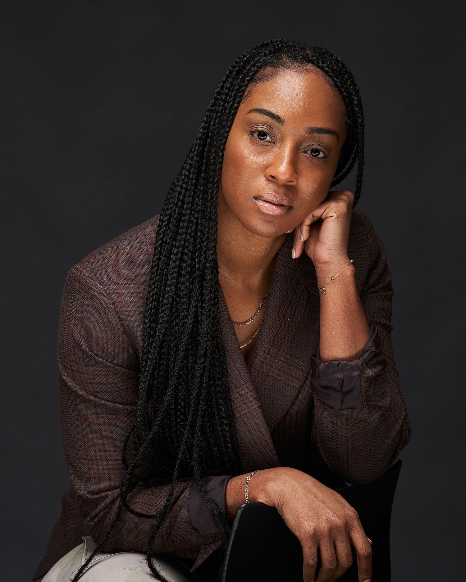 Woman with long box braids in brown blazer posing in thoughtful seated position against dark background.