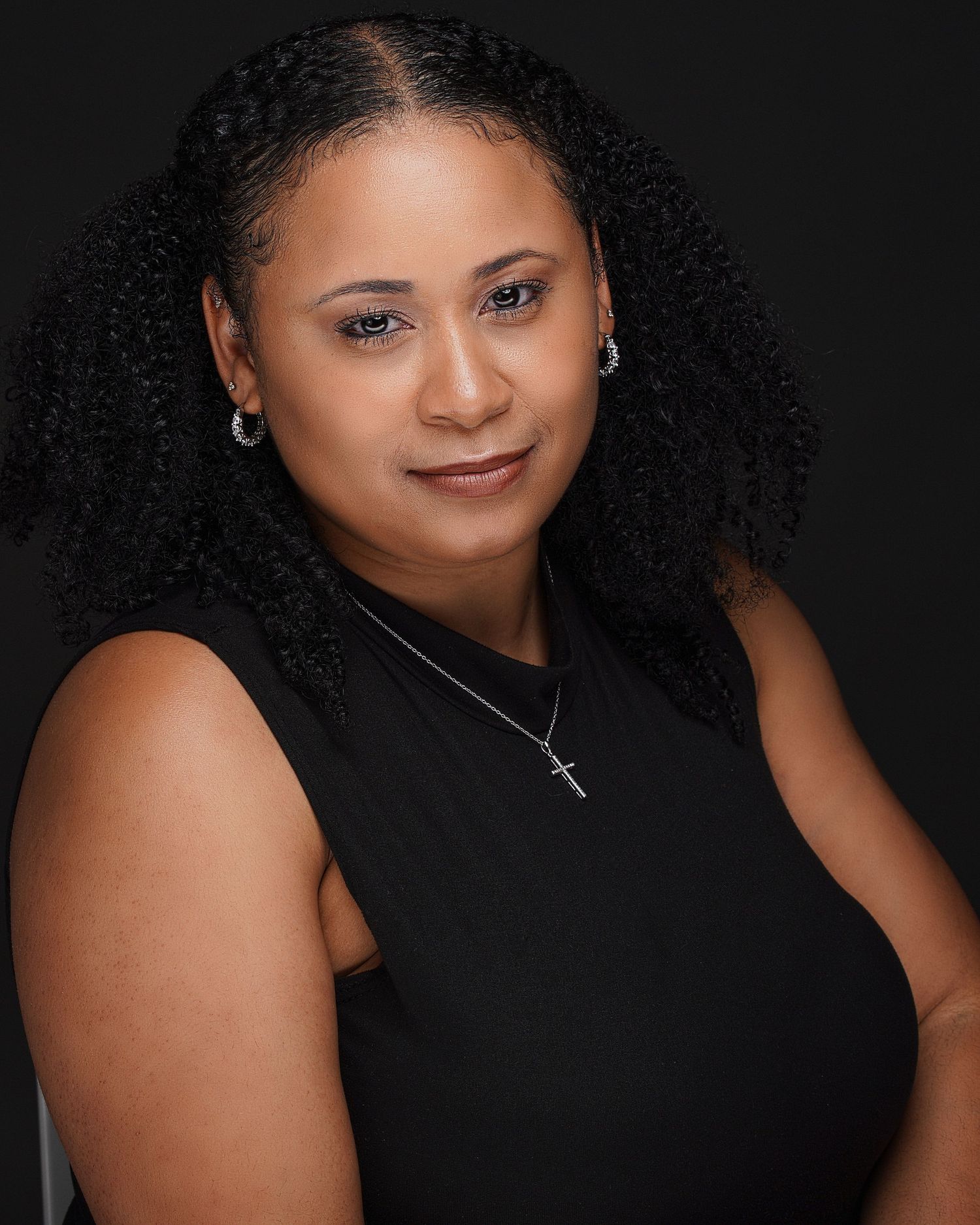 Professional woman with natural curly hair in black sleeveless top wearing cross necklace against dark background.