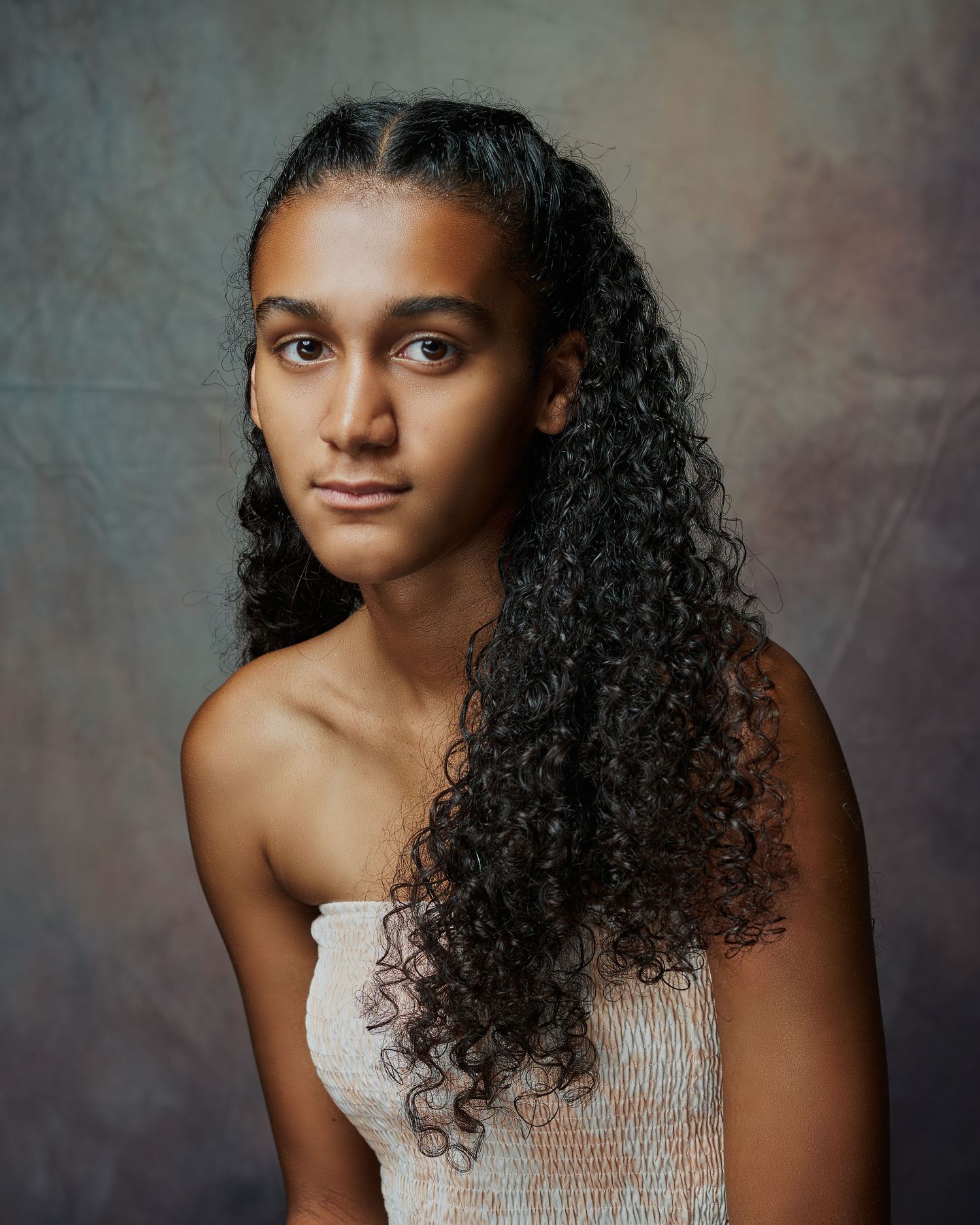 Young woman with long curly hair in white strapless top posing against textured studio background.