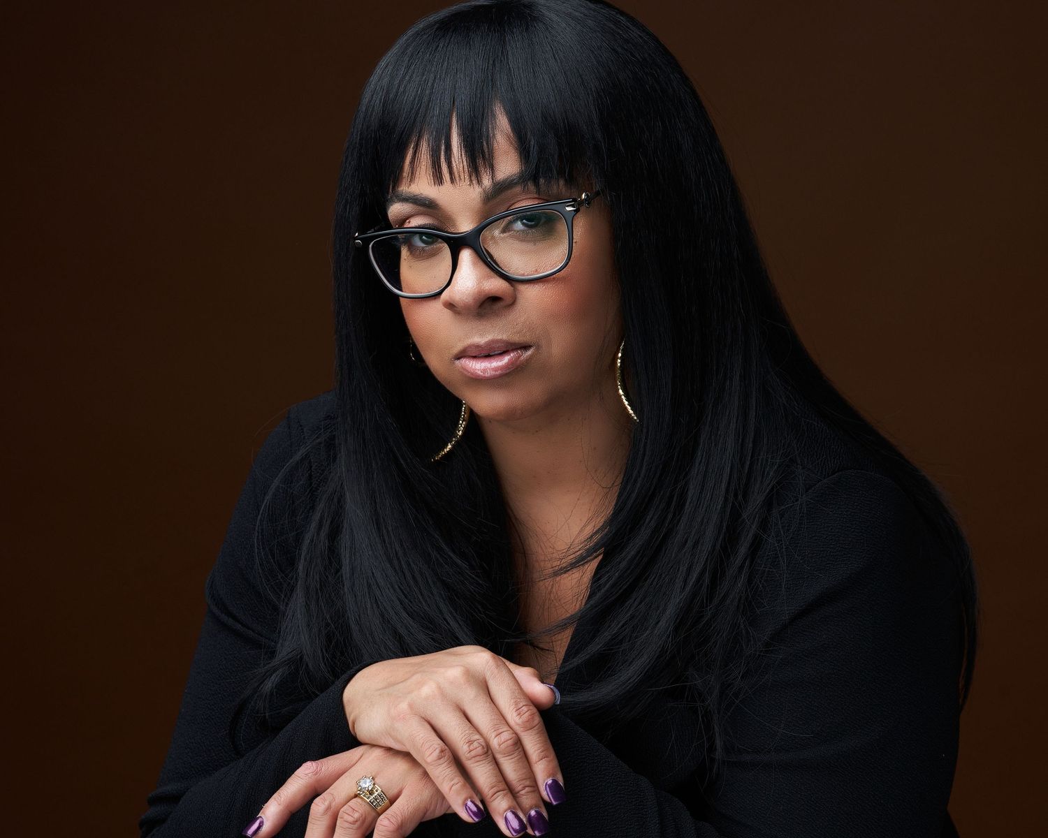 A woman with straight black hair and glasses poses confidently in an all-black outfit with gold hoop earrings.
