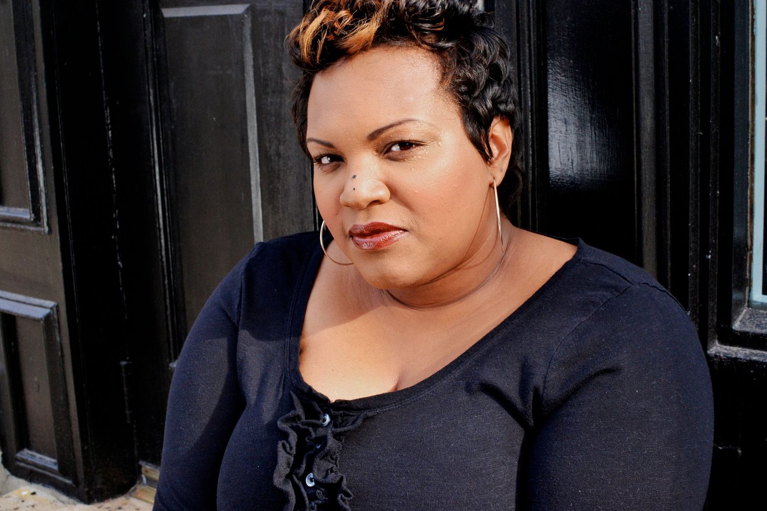 Plus-size Black woman with short hair wearing navy blue top seated against black door exterior.