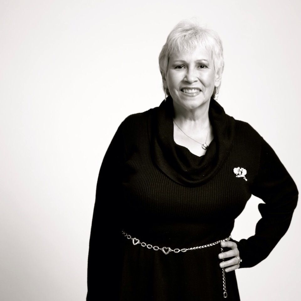 Black and white portrait of a smiling woman with short white hair wearing a black cowl-neck sweater with a chain belt.