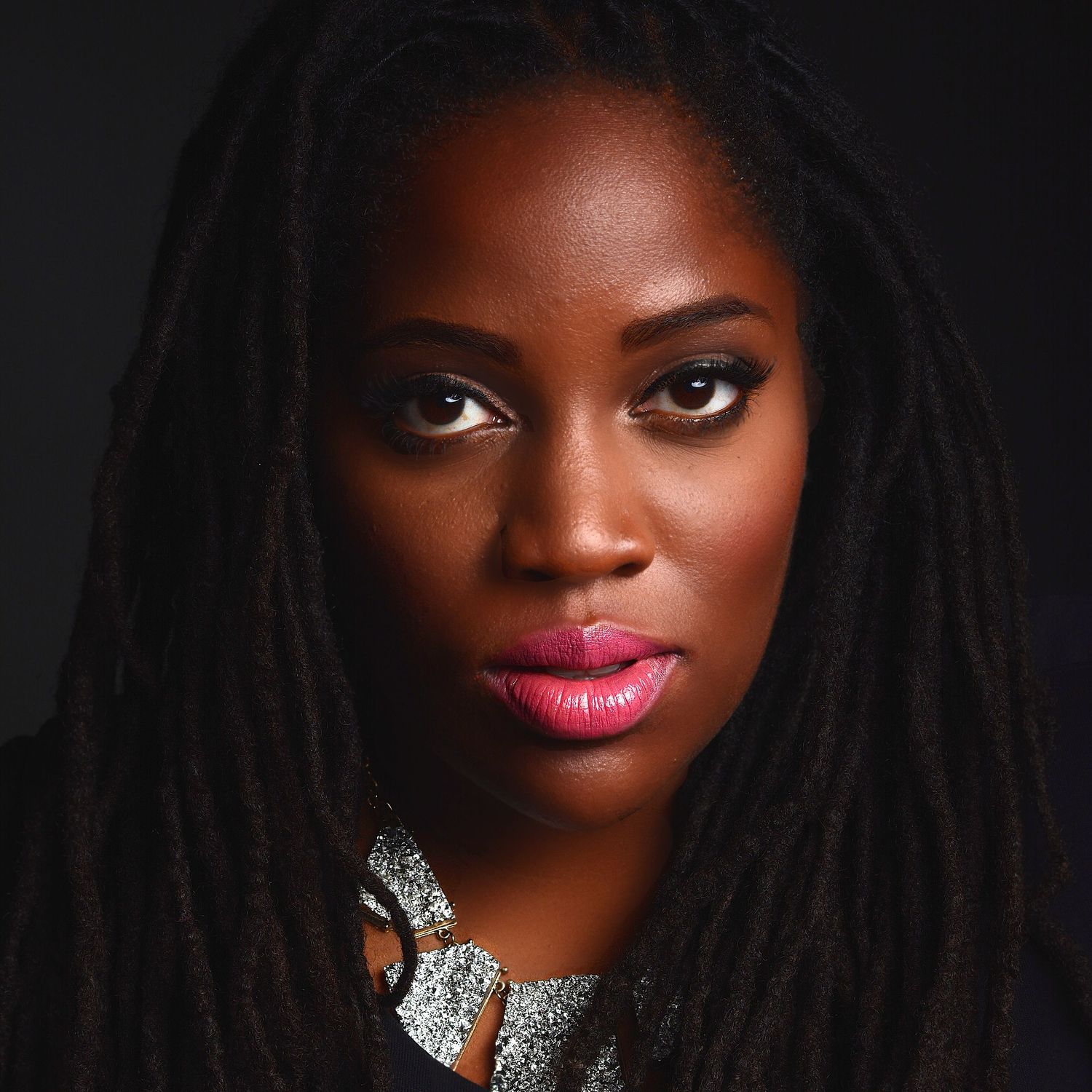 A young Black woman with long locs, bold pink lips, and a silver necklace poses against a dark background.