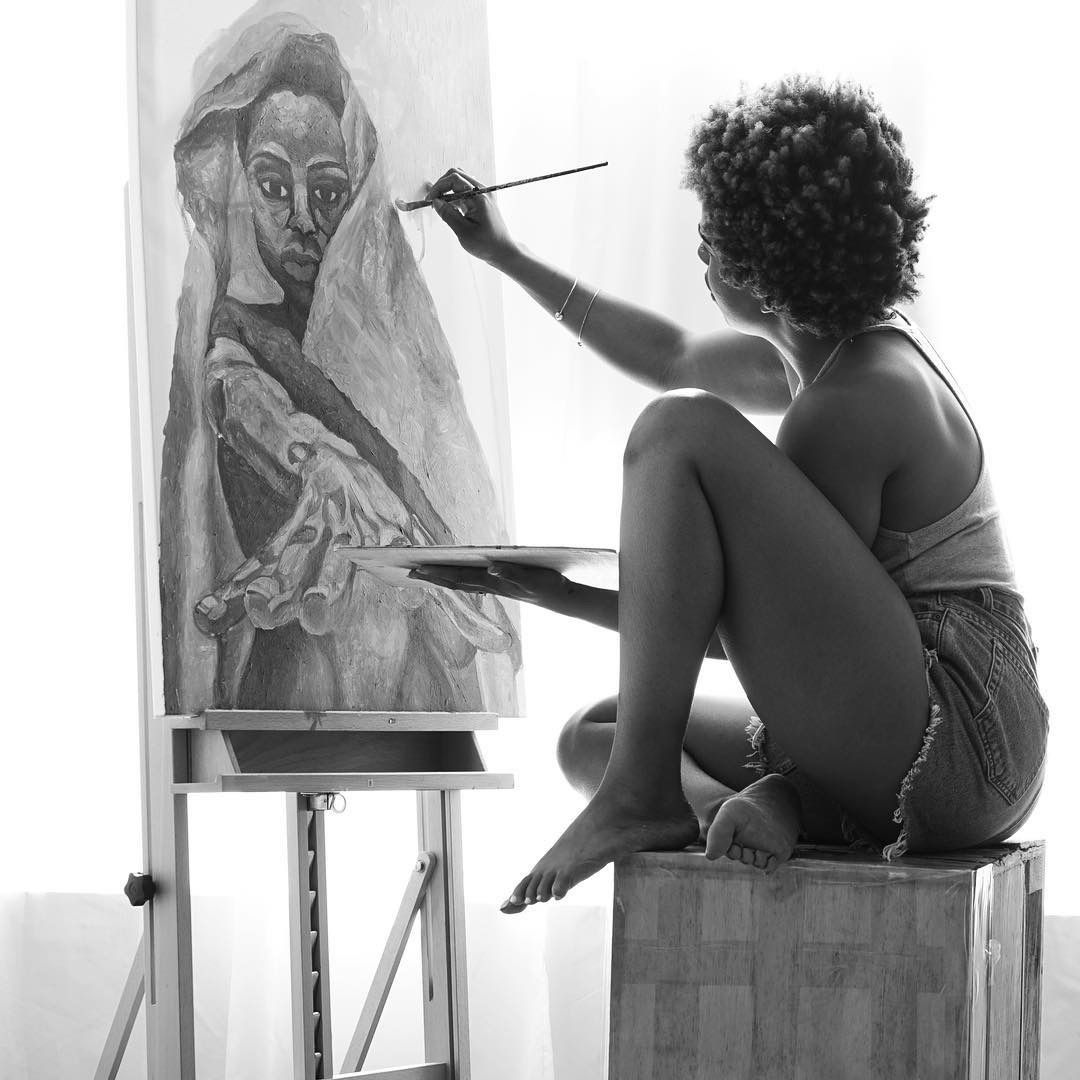 Black and white photo of a woman painting a portrait on canvas while seated on a wooden block.