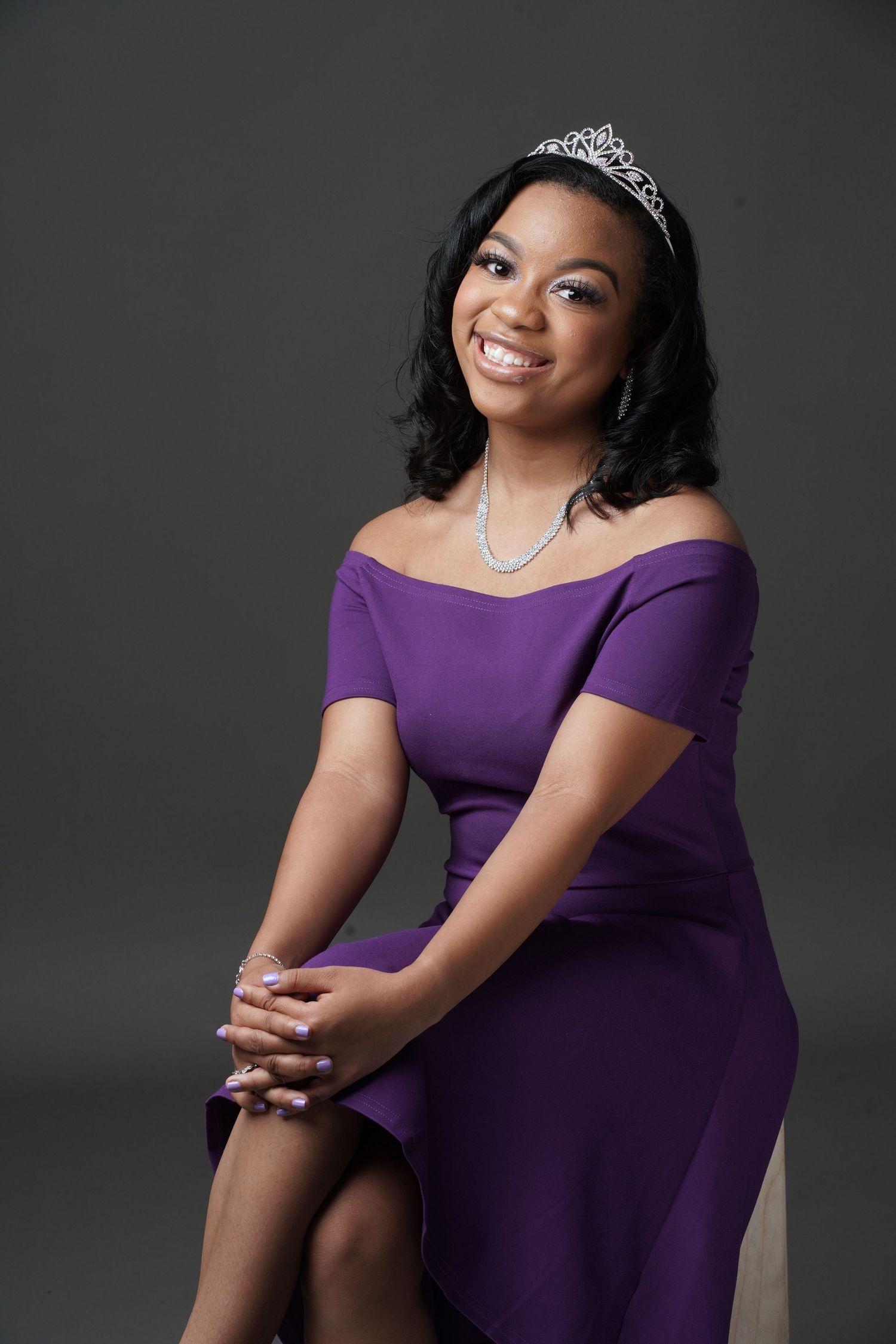 Young woman in purple off-shoulder dress wearing tiara and pearl necklace seated.