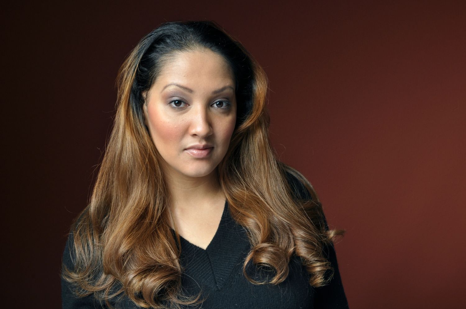 A woman with long highlighted hair poses confidently against a deep red background in a black top.