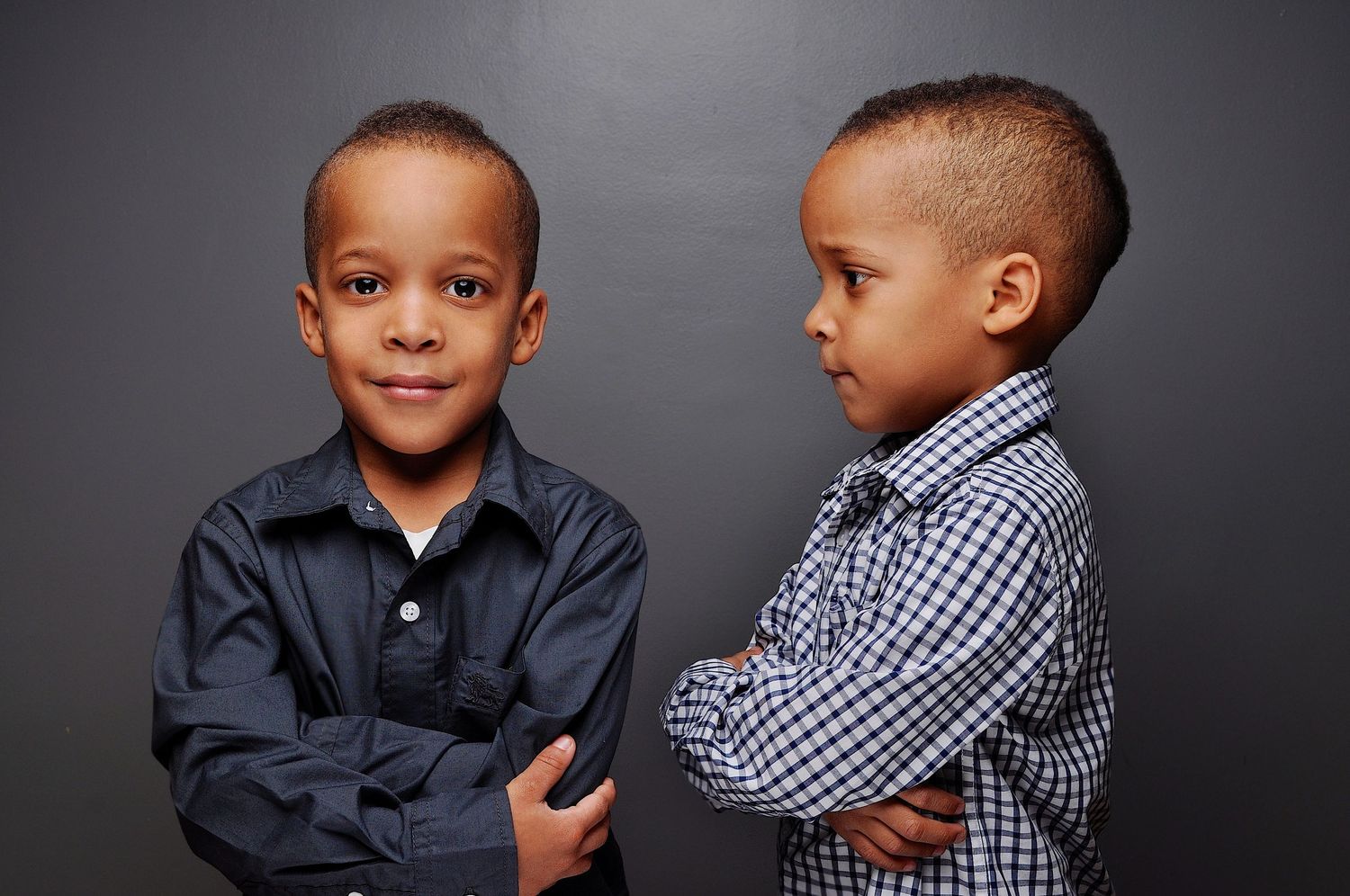 Two young boys in button-up shirts stand with arms crossed against a gray background.