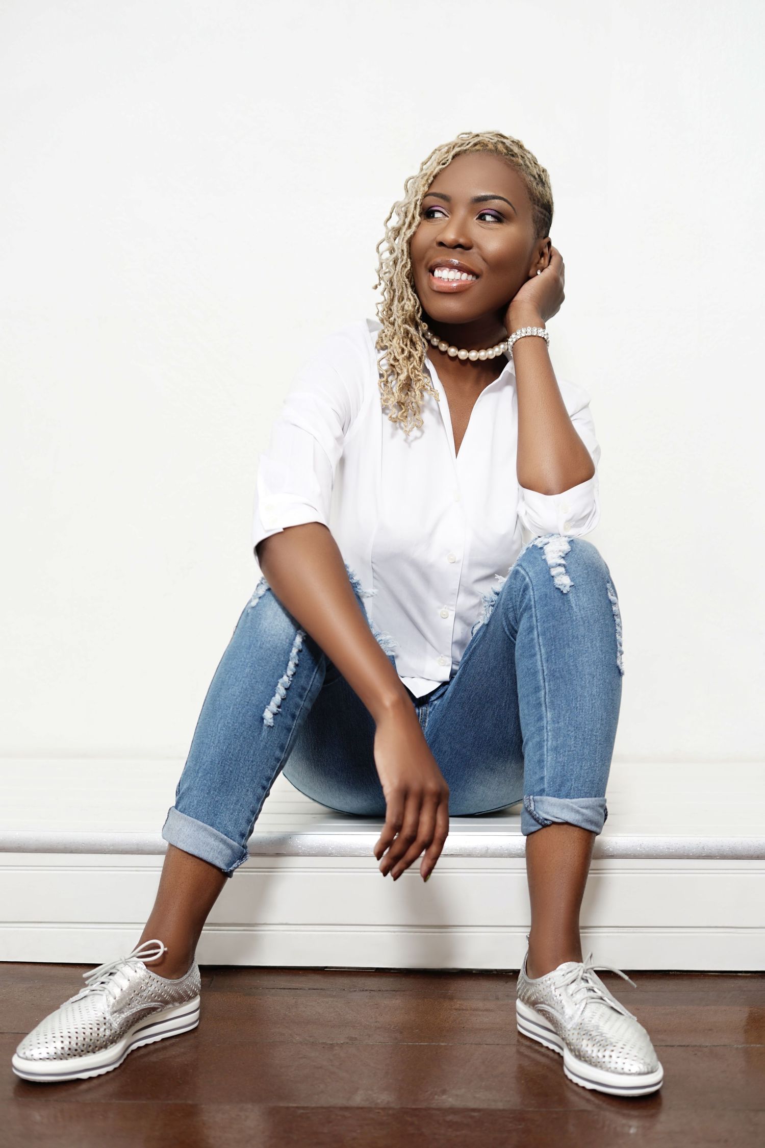A smiling Black woman with blonde curly hair sits casually in ripped jeans and white shirt against a white wall.